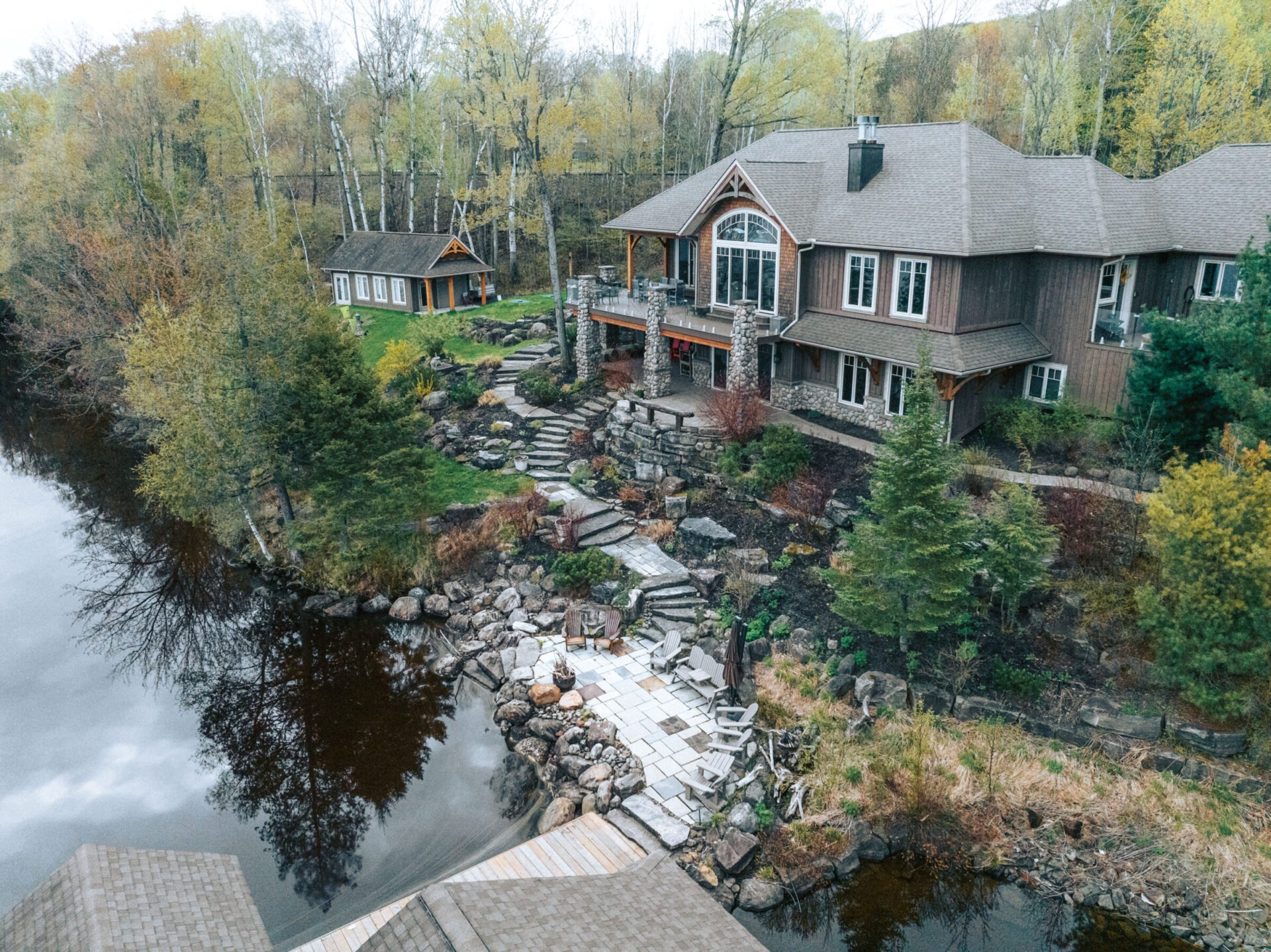 Aerial view of a large house beside a river, surrounded by trees and landscaped gardens, featuring a stone patio and pathway.