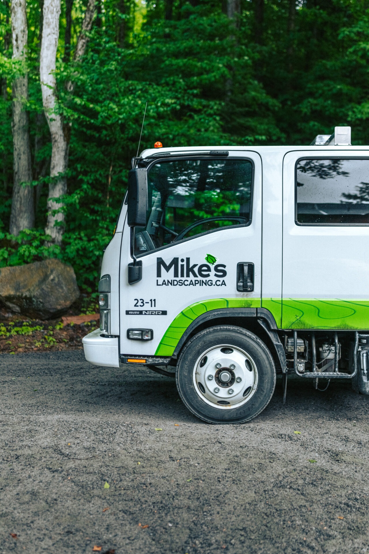 A white truck with "Mike's Landscaping" logo is parked on a gravel path surrounded by lush green forest trees.
