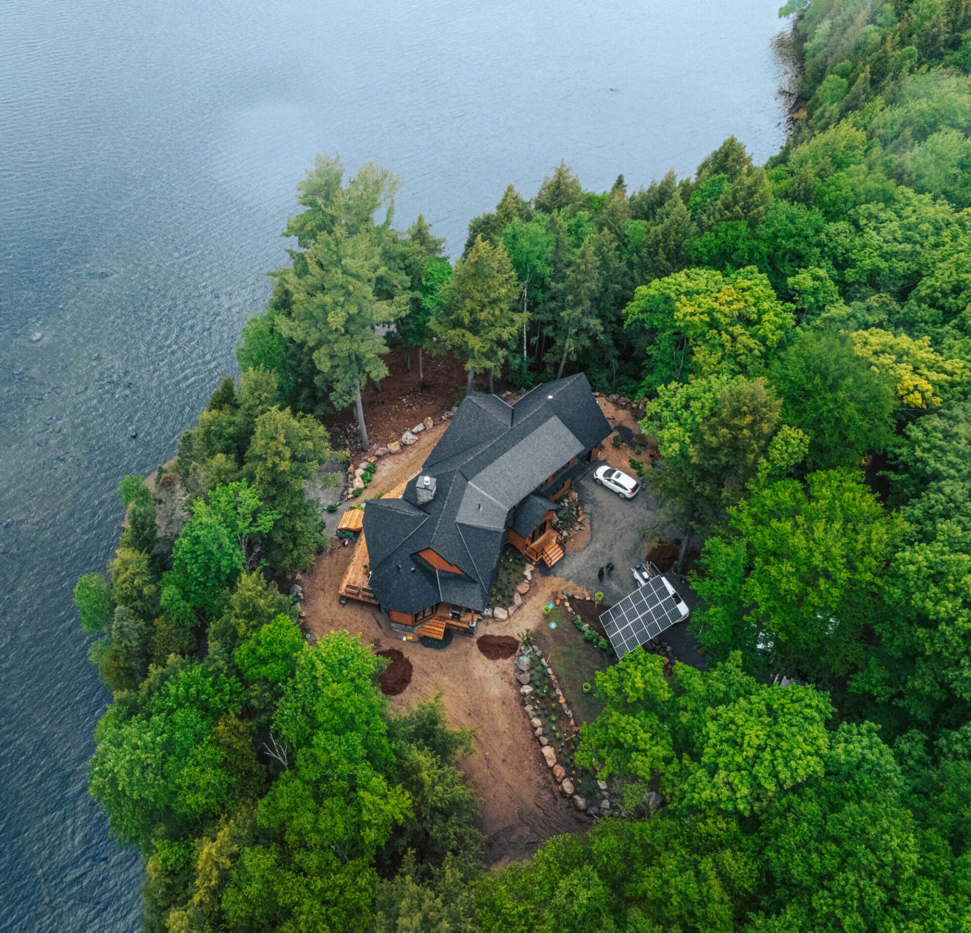 Aerial view of a lakeside house surrounded by lush forest, featuring a vehicle, solar panels, and a clear, calm body of water.