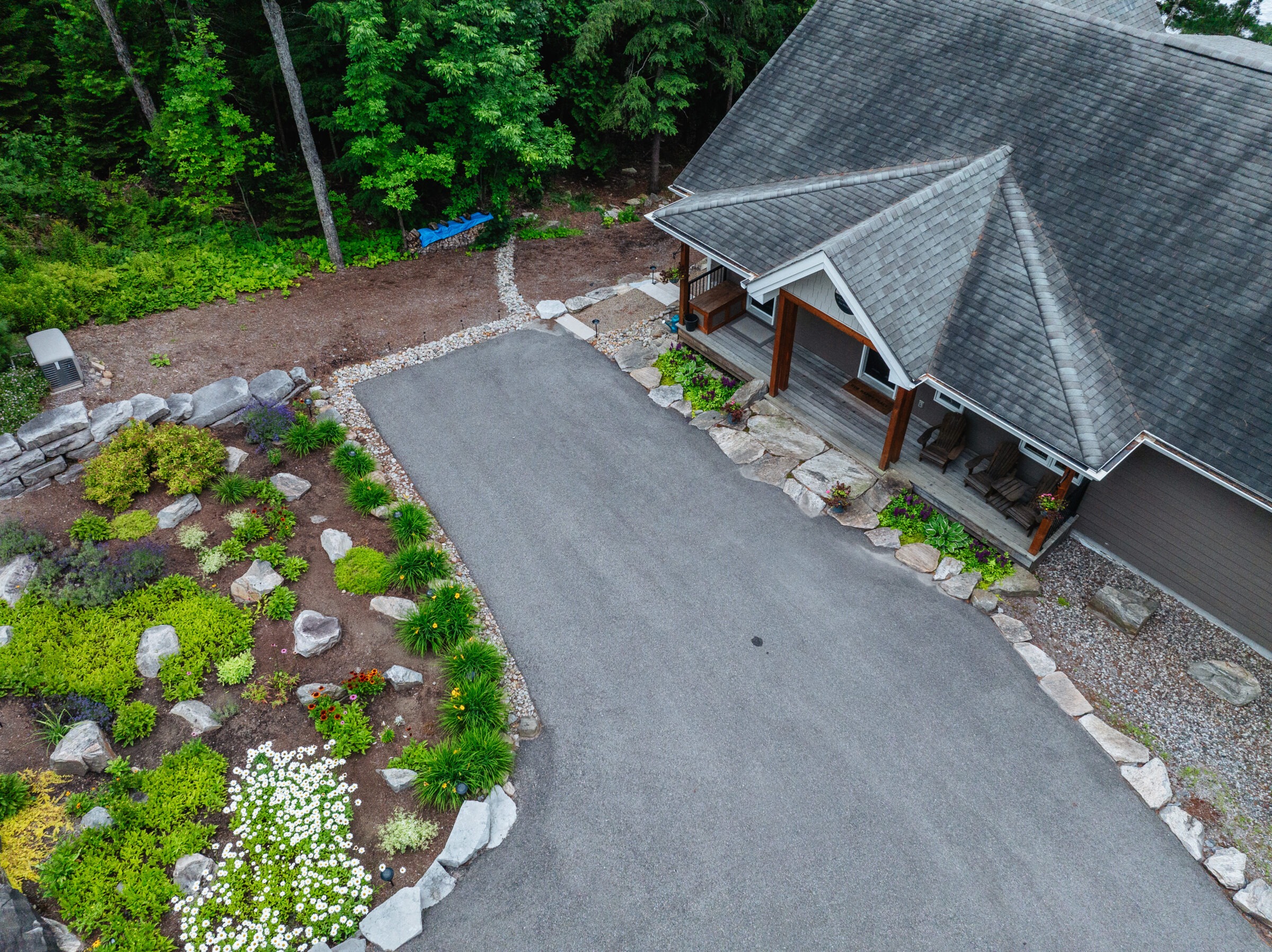 Aerial view of a house with a gray roof, surrounded by landscaped garden with rocks, plants, and trees. No people are visible.