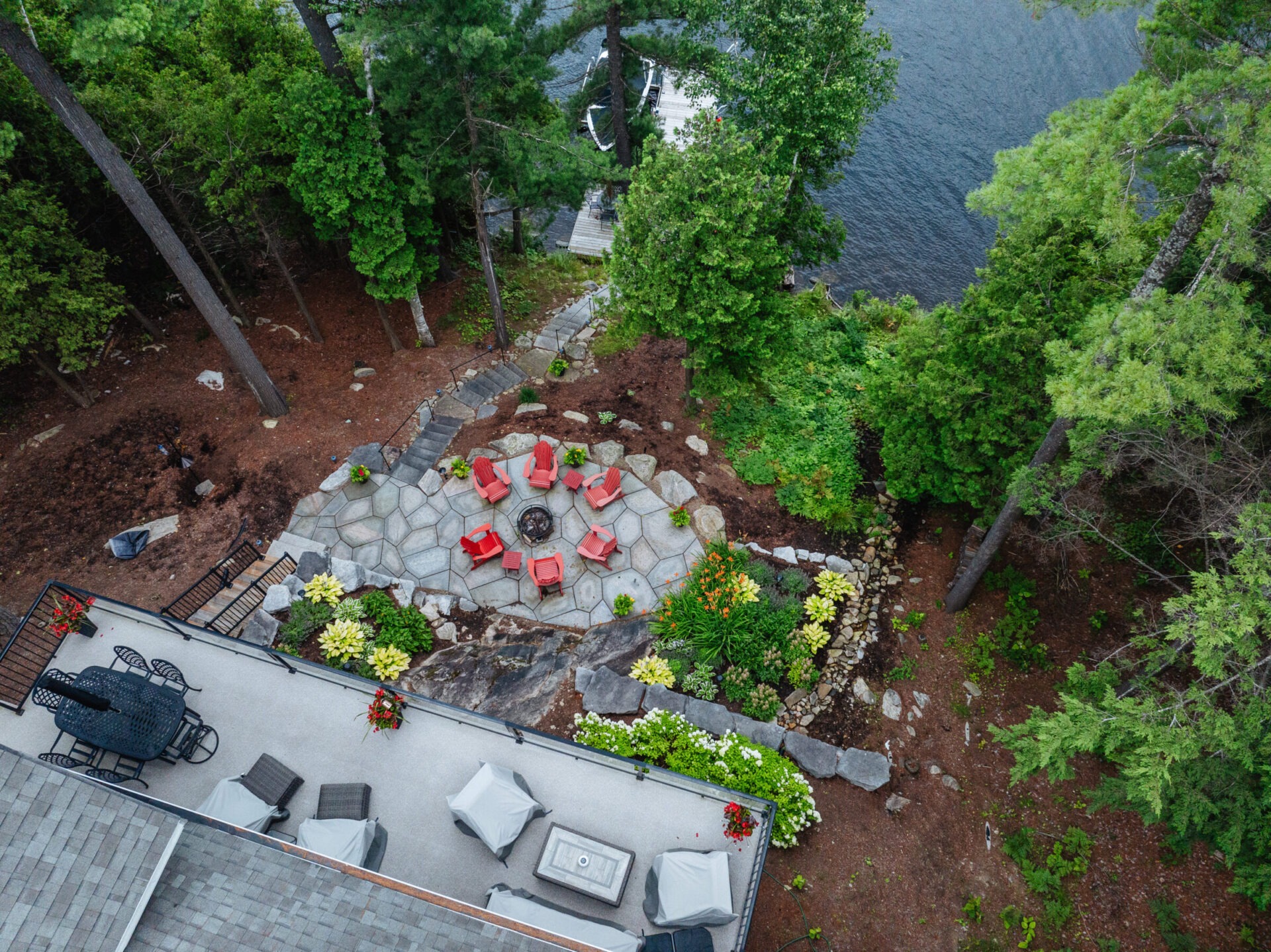 Aerial view of a serene lakeside patio with red chairs around a fire pit, surrounded by lush greenery and docks.
