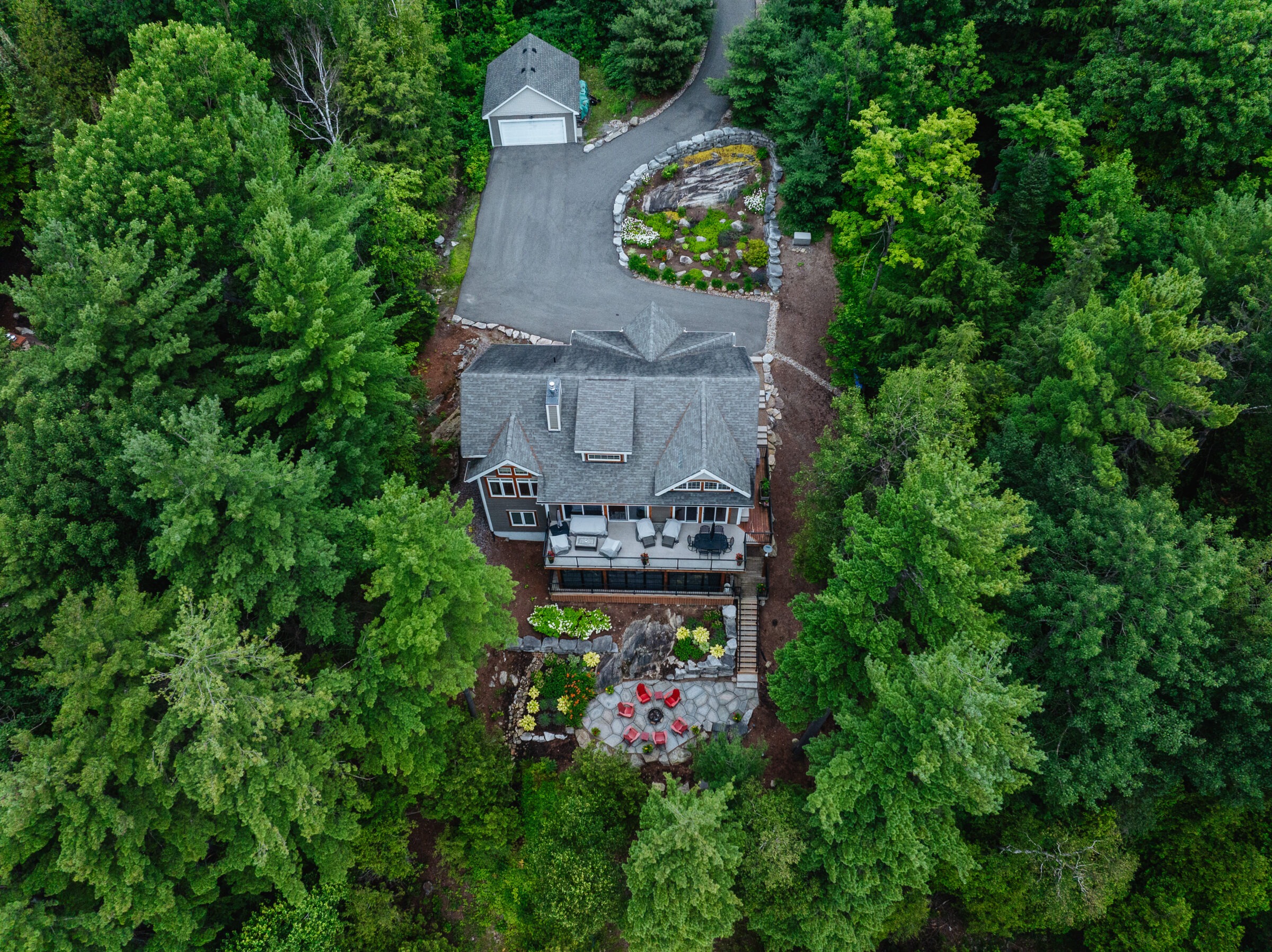 Aerial view of a house surrounded by lush green trees, with a driveway, patio, and garden visible in a forest setting.