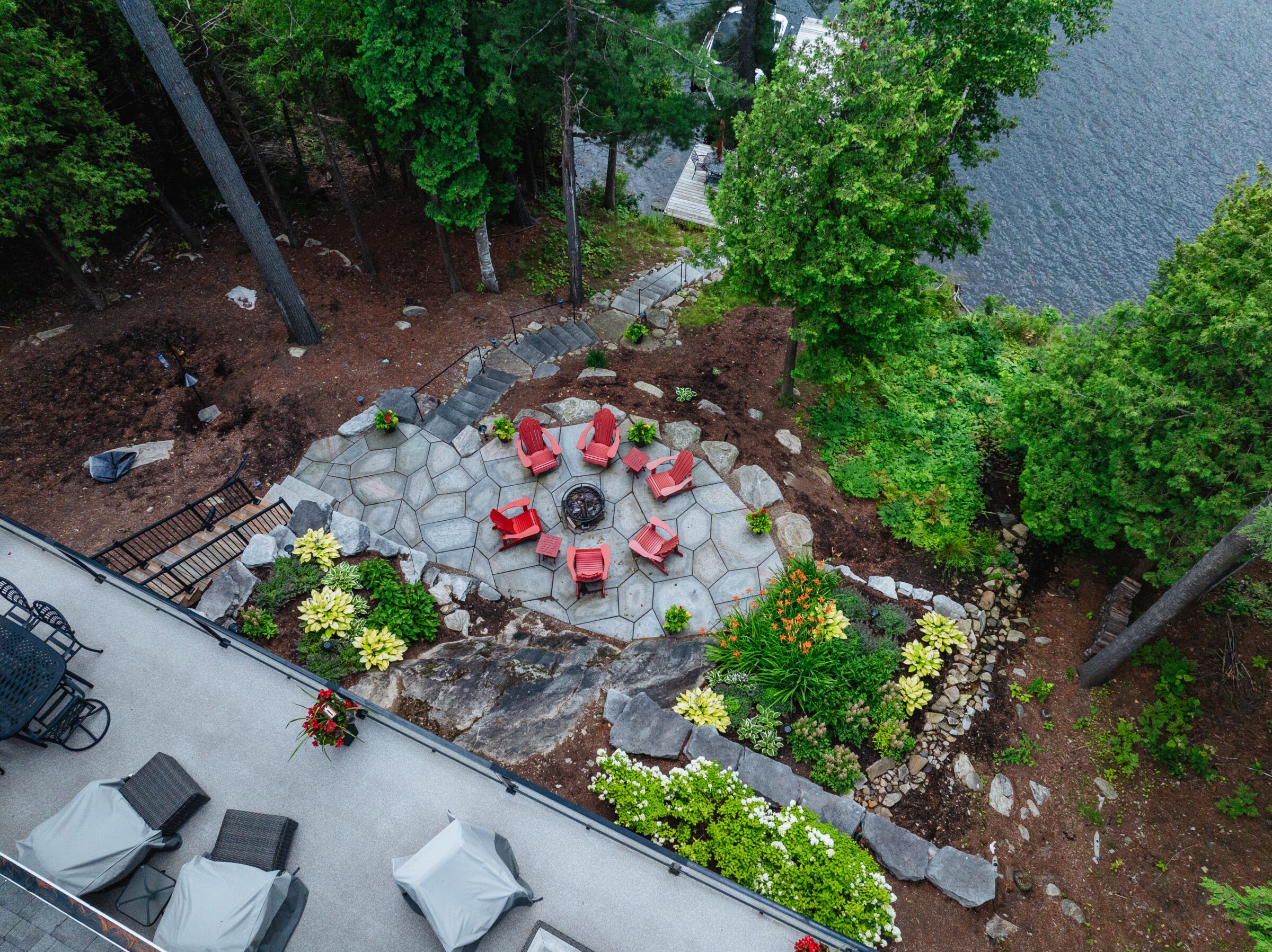 Stone patio with red chairs arranged around a fire pit, surrounded by lush trees and overlooking a serene body of water.