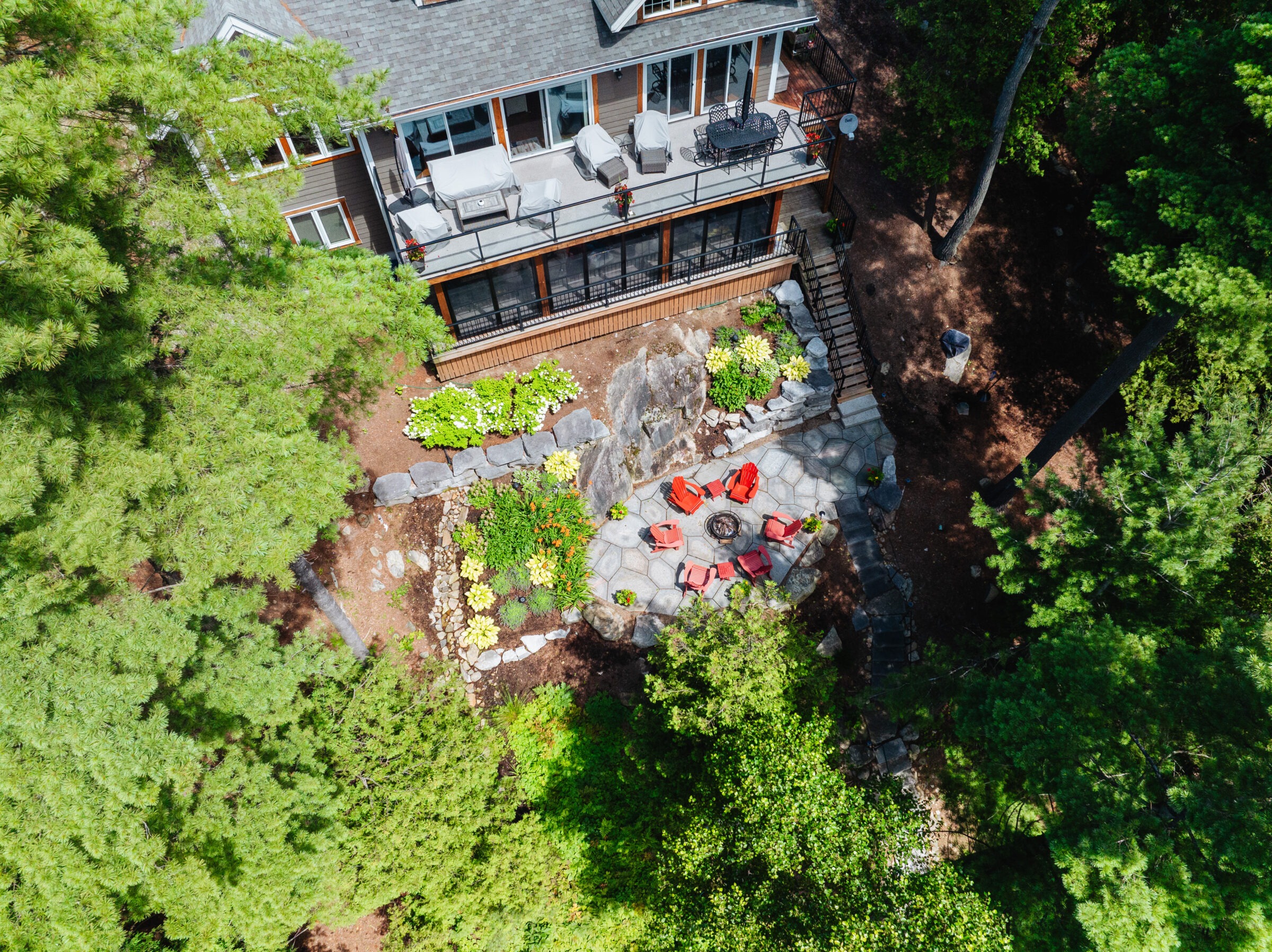 Aerial view of a forest-surrounded house with a patio featuring red chairs and a fire pit, lush greenery around.