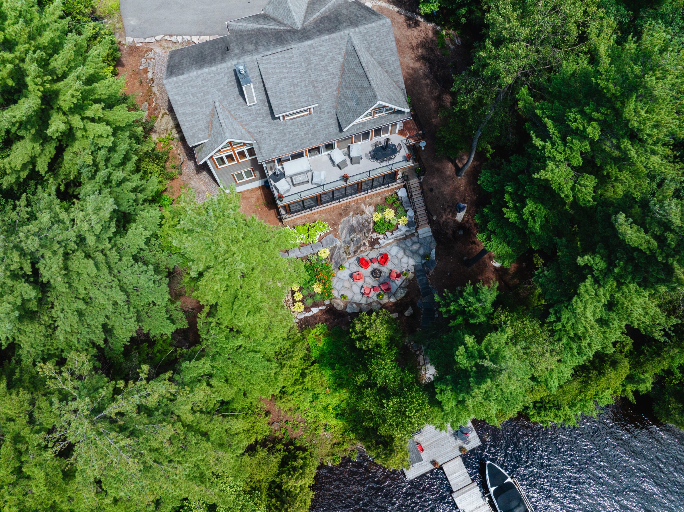 Aerial view of a lakeside house surrounded by lush trees, featuring a patio with red chairs and a dock with a boat.