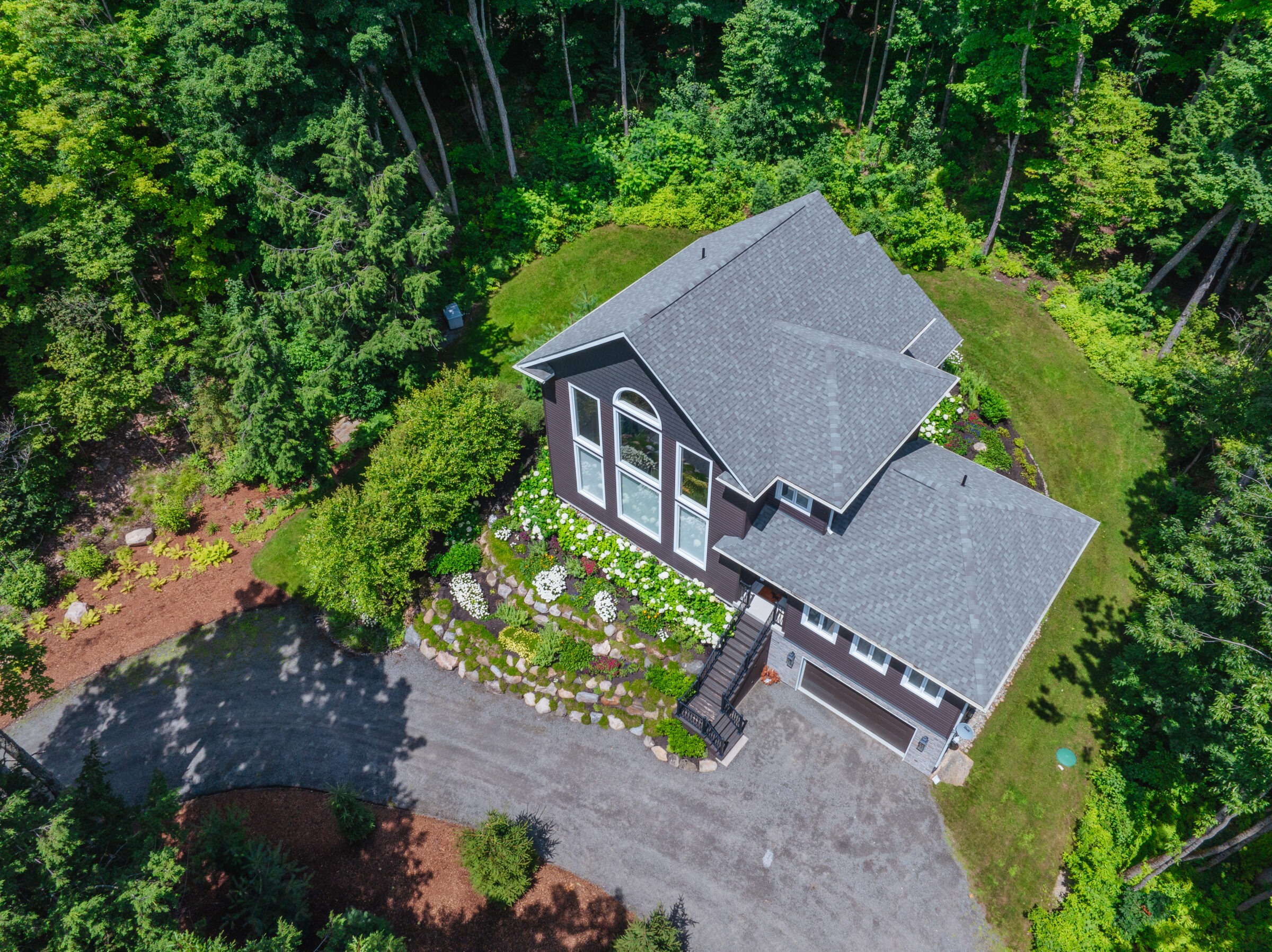 Aerial view of a modern house surrounded by lush greenery, landscaped garden, and winding driveway amidst dense forest.