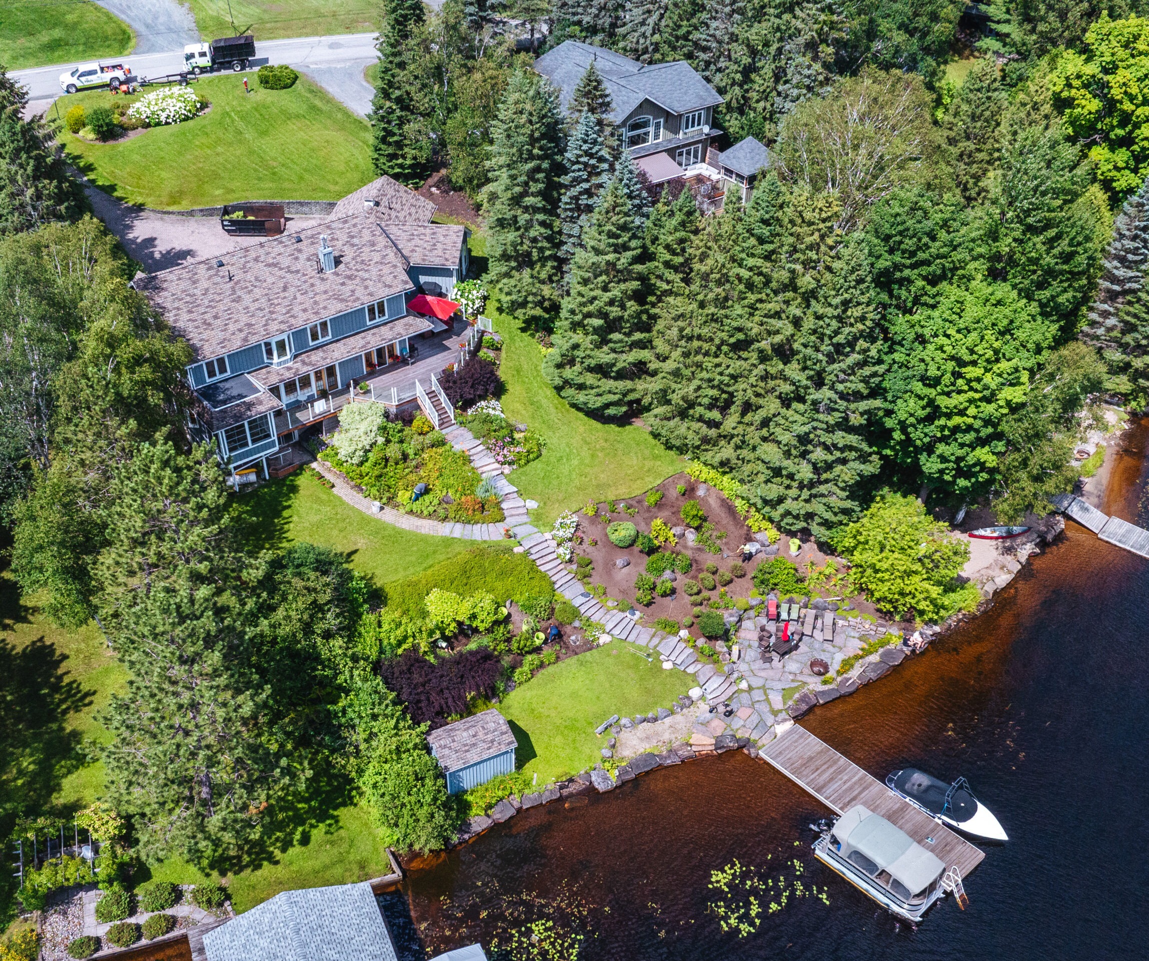 Aerial view of a lakeside property with two houses, lush gardens, a dock with boats, surrounded by trees and a road nearby.