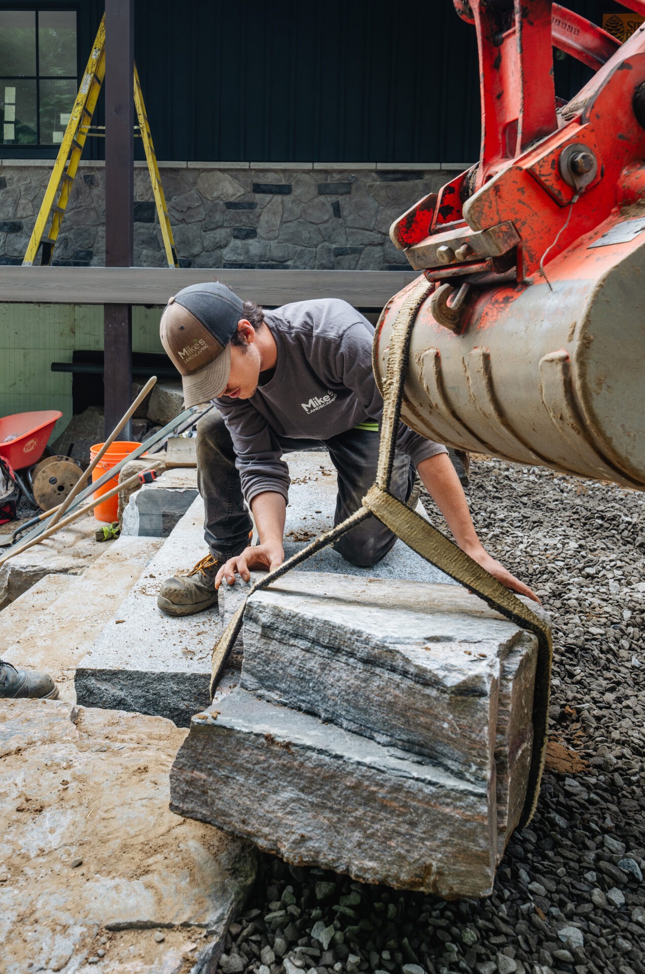 A person is guiding a large stone slab with a machine’s help at a construction site. Tools and ladders are visible.
