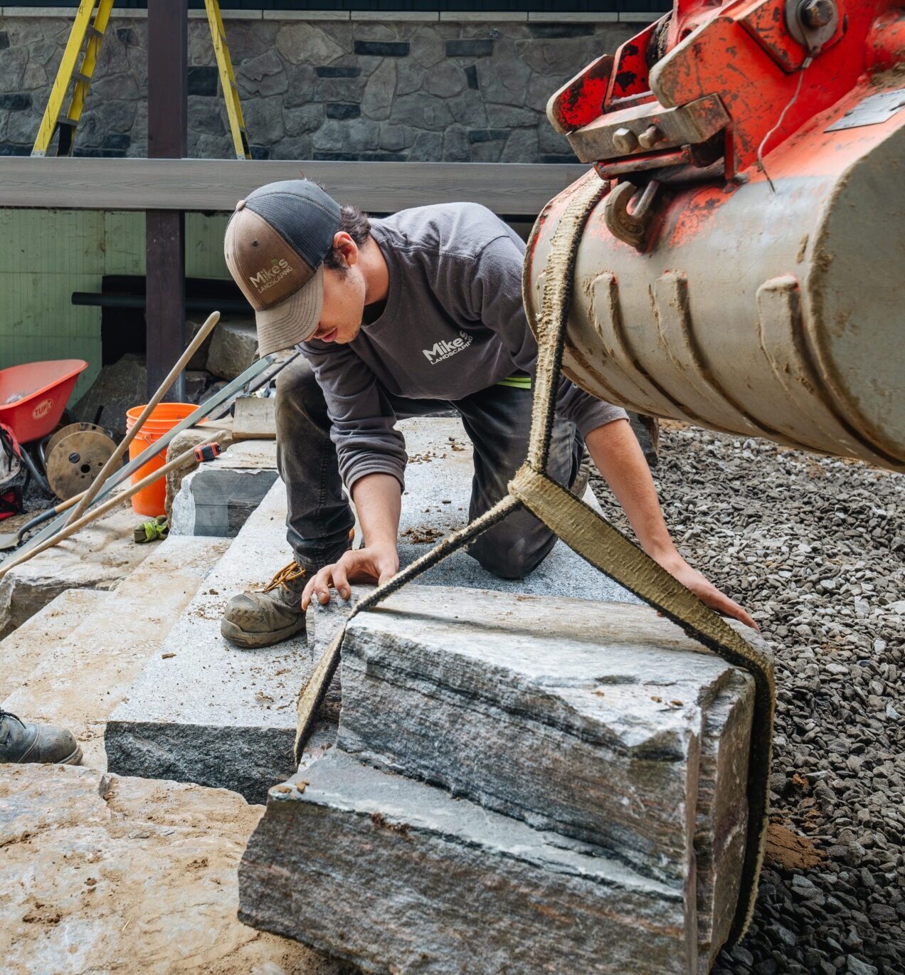 A person works with large stones using a lifting mechanism and tools, wearing workwear in an outdoor construction setting with stone walls.