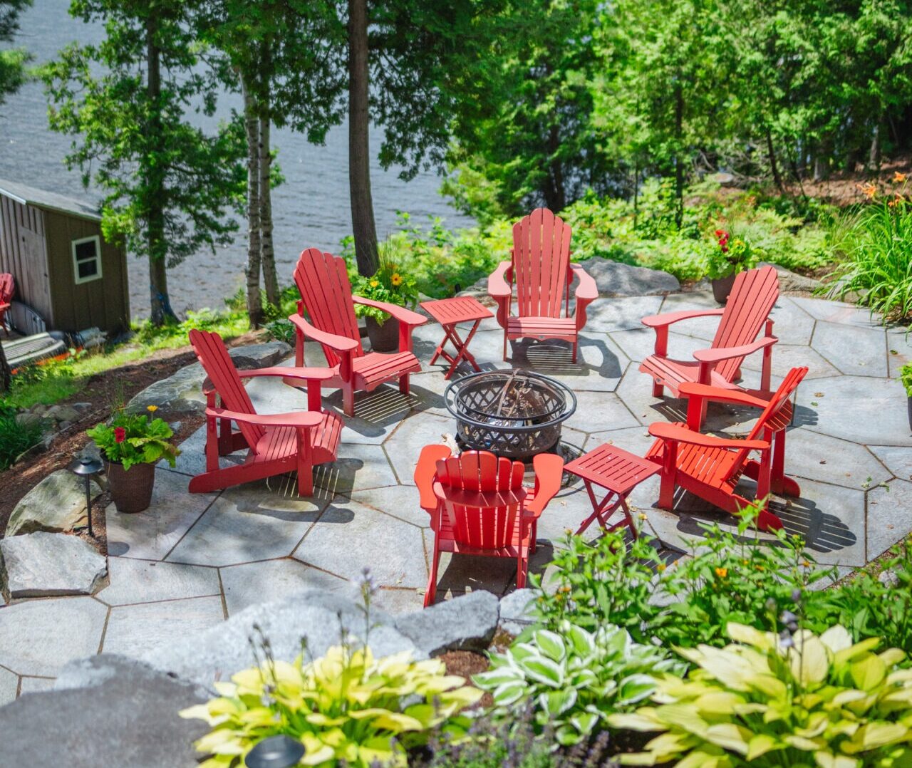 Red Adirondack chairs encircle a fire pit on a stone patio, surrounded by lush greenery and overlooking a tranquil lake, creating a serene ambiance.