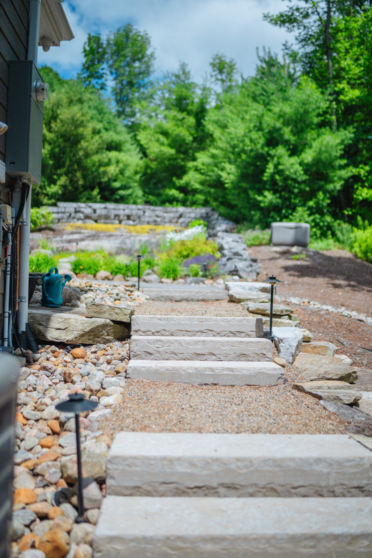 Stone steps lead through a lush garden, surrounded by greenery and rock landscaping, under a bright, partly cloudy sky.