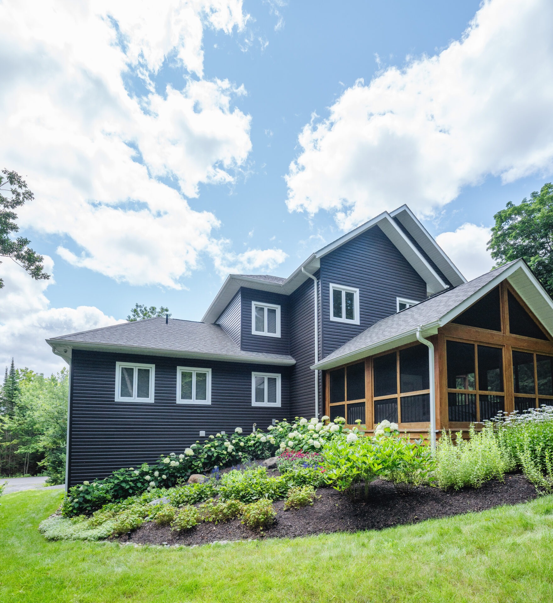A modern house with dark siding and large windows, surrounded by lush landscaping and clear blue skies above, showcasing a serene setting.