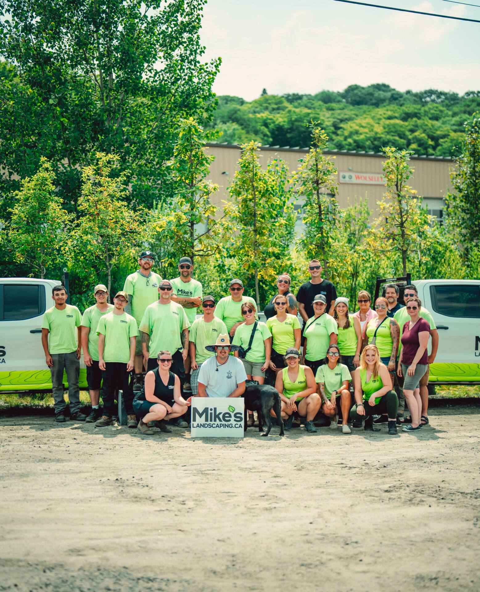 A group of people in matching shirts poses outdoors with a landscaping truck and sign, surrounded by green trees and a building.
