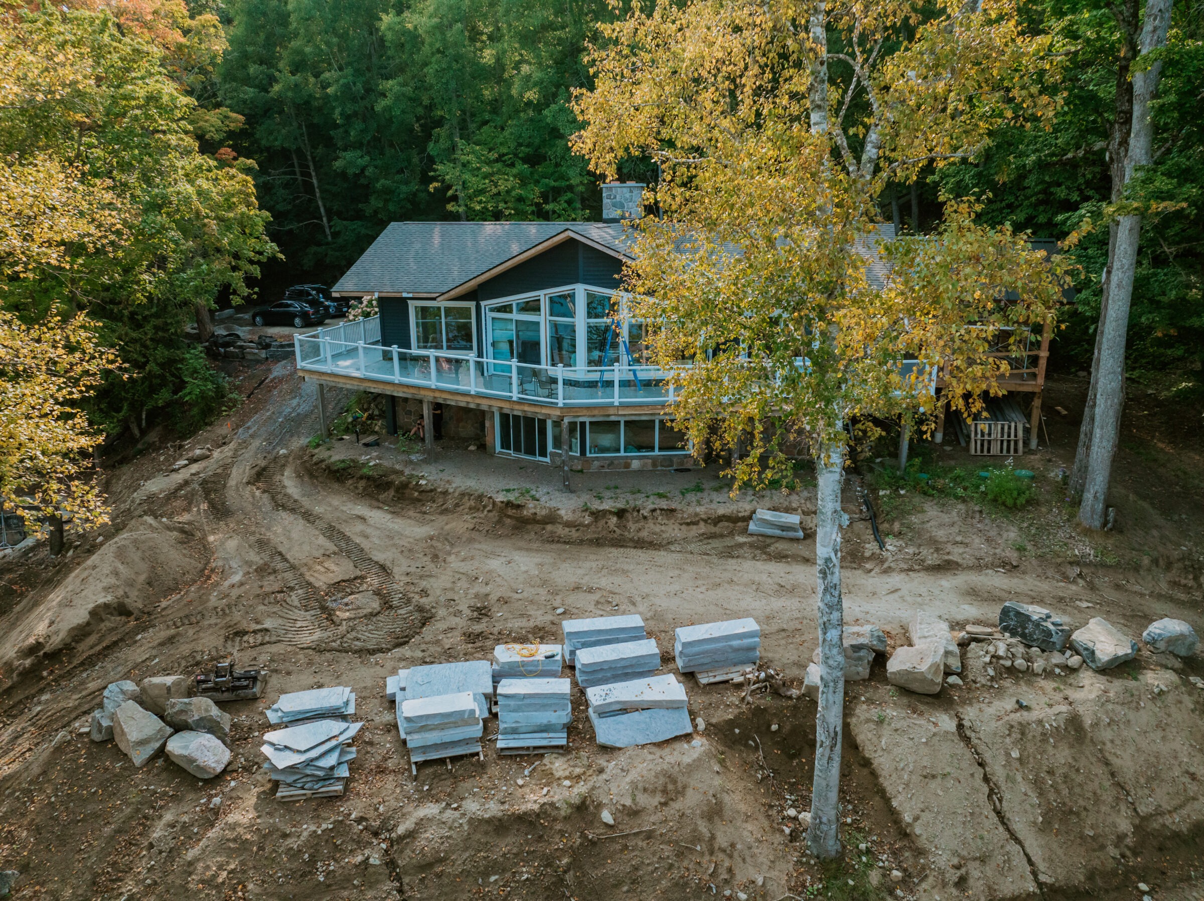 A modern house with floor-to-ceiling windows amidst trees, surrounded by construction materials and cleared land.