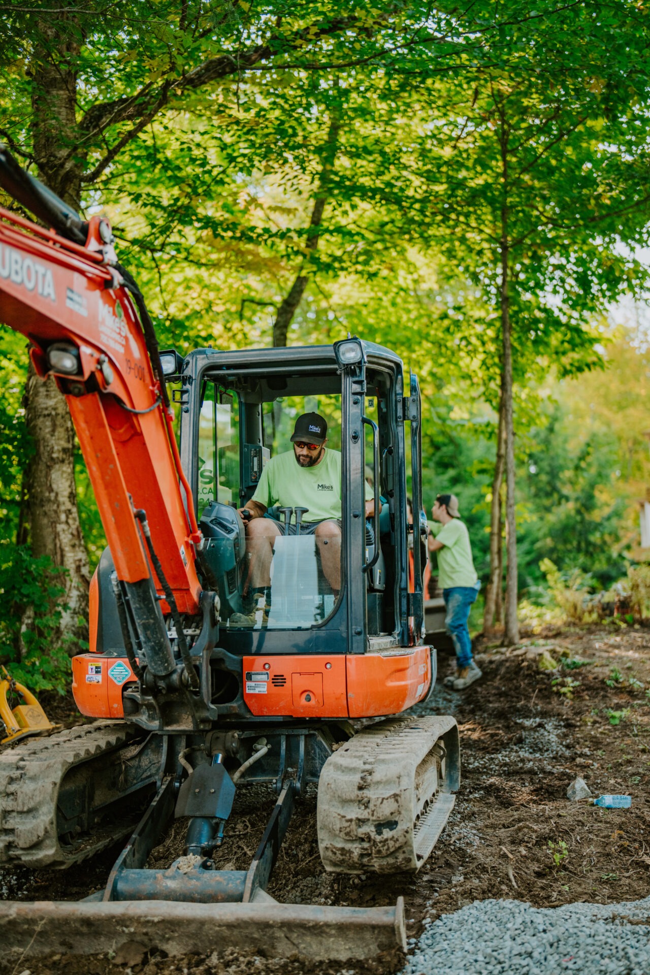A person operates an orange excavator in a lush, wooded area. Another person stands nearby. No recognizable landmarks or historical buildings present.