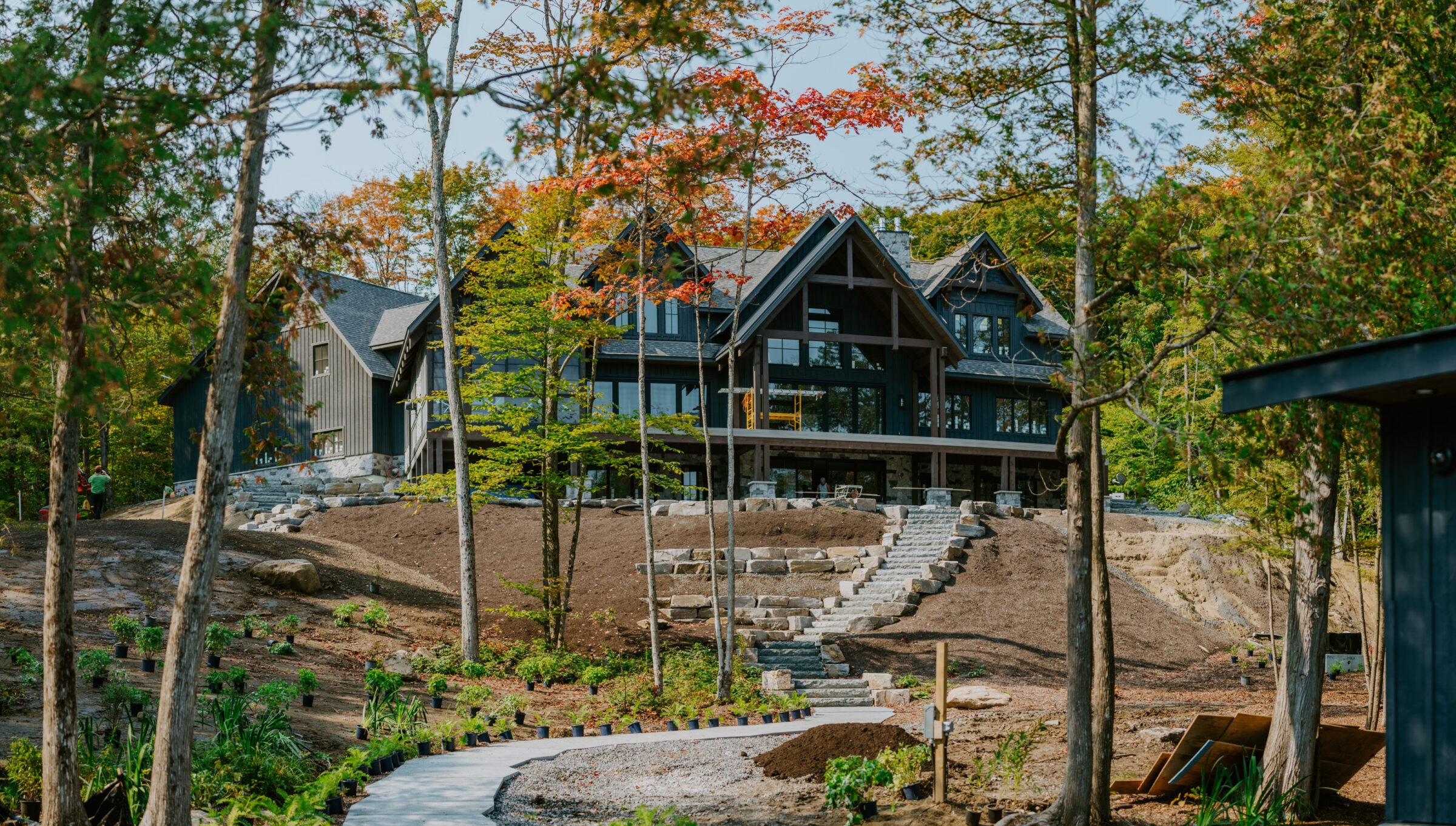 A large, modern cabin surrounded by vibrant autumn trees and a landscaped garden, with a person working near the path leading to it.