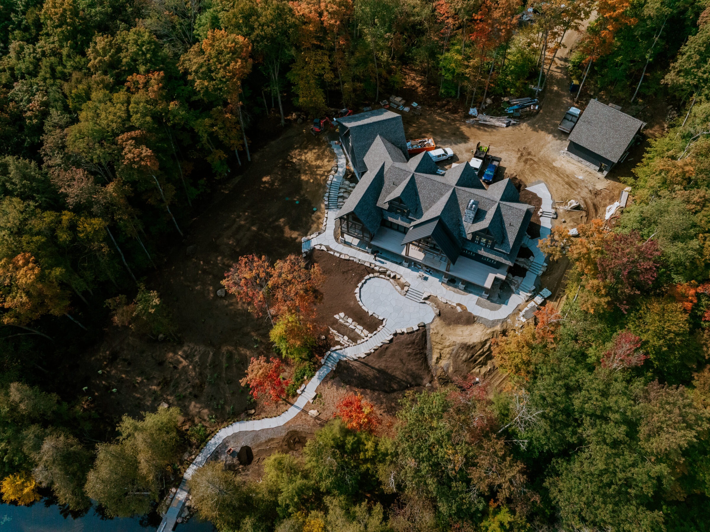 Aerial view of a large house surrounded by dense forest, featuring winding pathways and autumn foliage, with construction vehicles nearby.