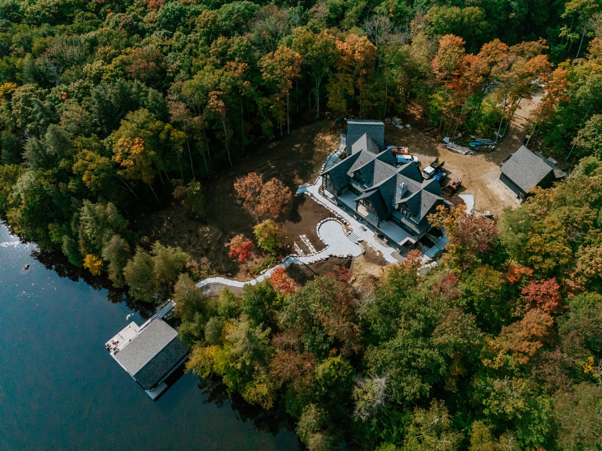 Aerial view of a large house surrounded by dense forest, colorful autumn foliage, and a nearby lake with a docked boathouse.