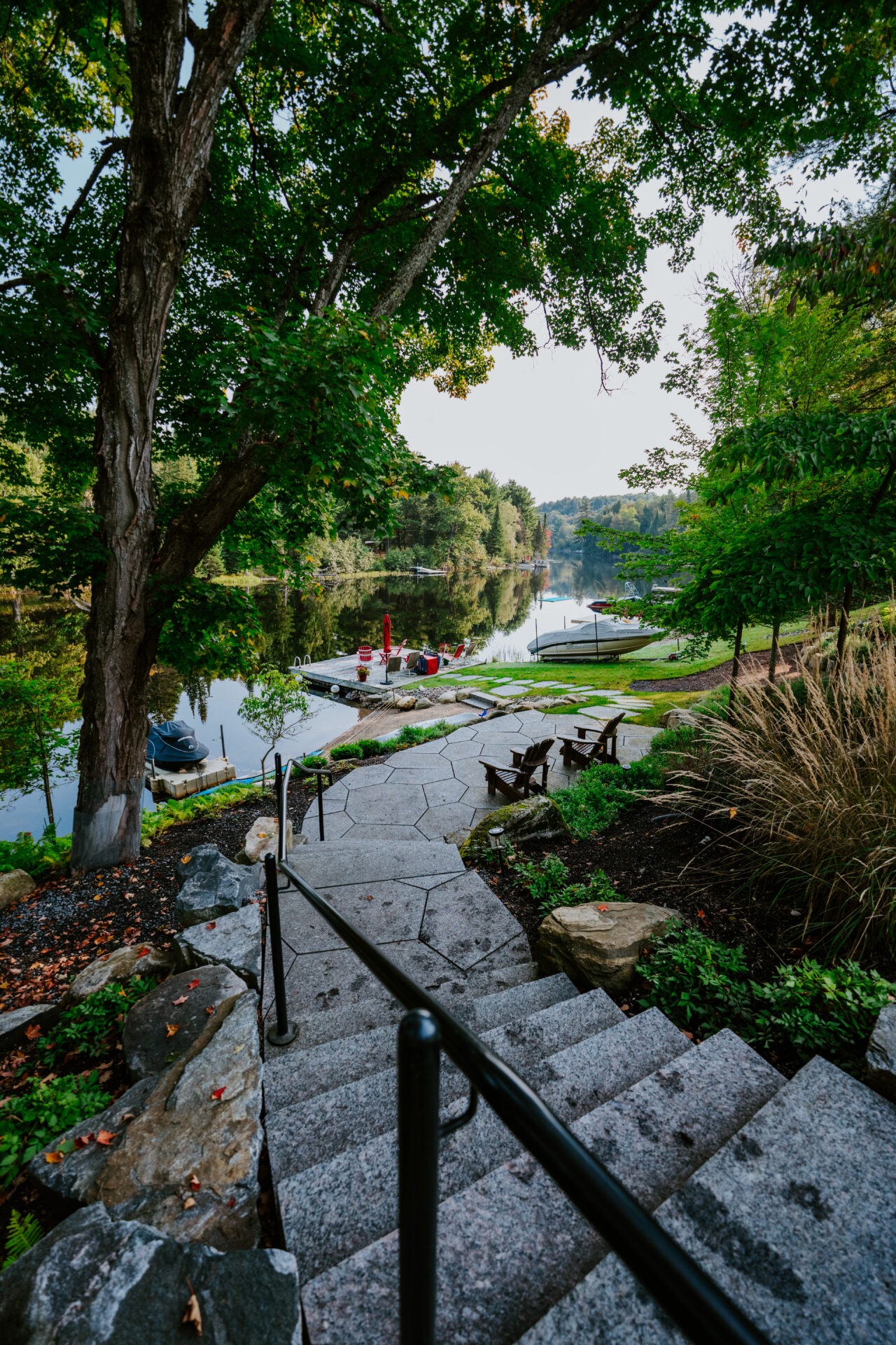 Stone steps descend through lush trees to a serene lakeside, with boats docked and chairs set for relaxation near the water.