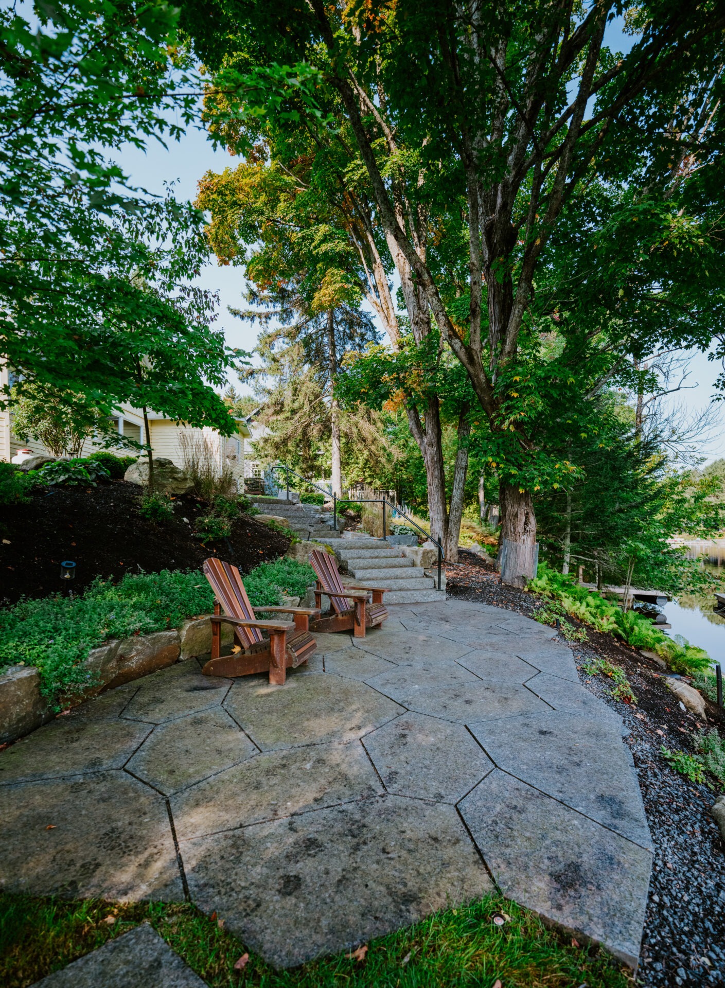 Two wooden chairs on a stone patio surrounded by lush green foliage and towering trees, near a peaceful riverbank.