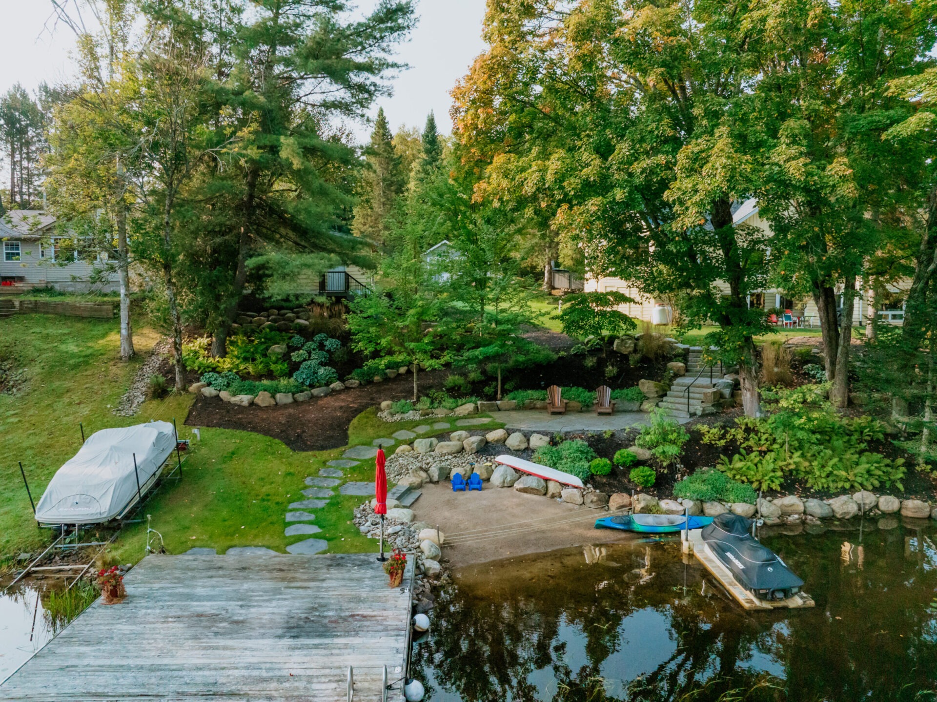 A picturesque lakefront garden features stone paths, a dock, seating area, and watercraft, surrounded by lush greenery and trees.