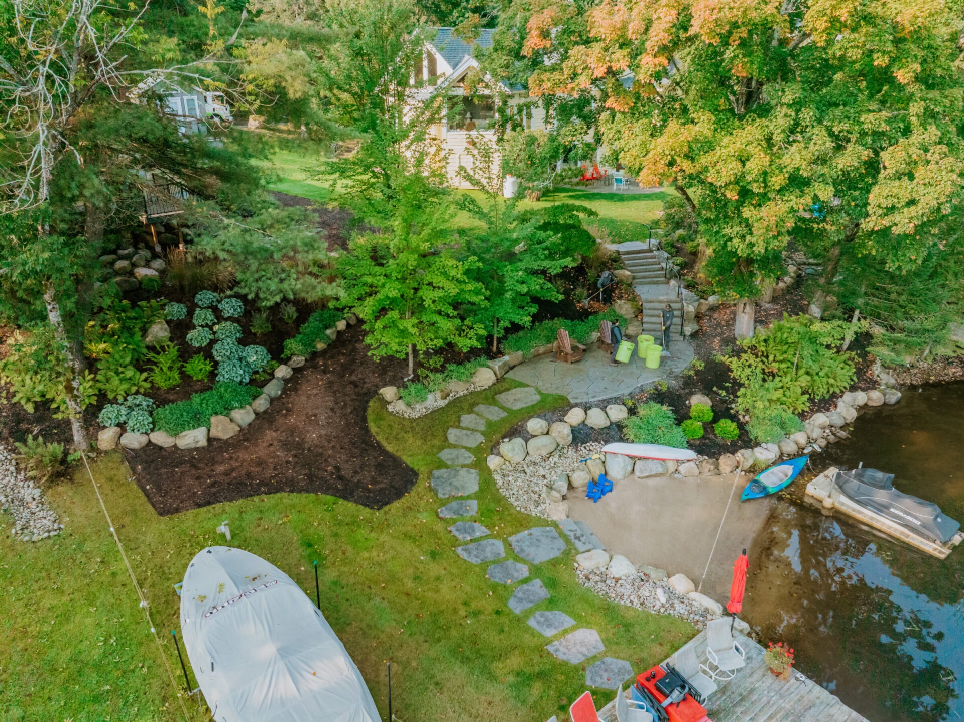 A scenic lakeside garden with stone pathways, chairs, and kayaks, surrounded by lush greenery and a calm water vista.