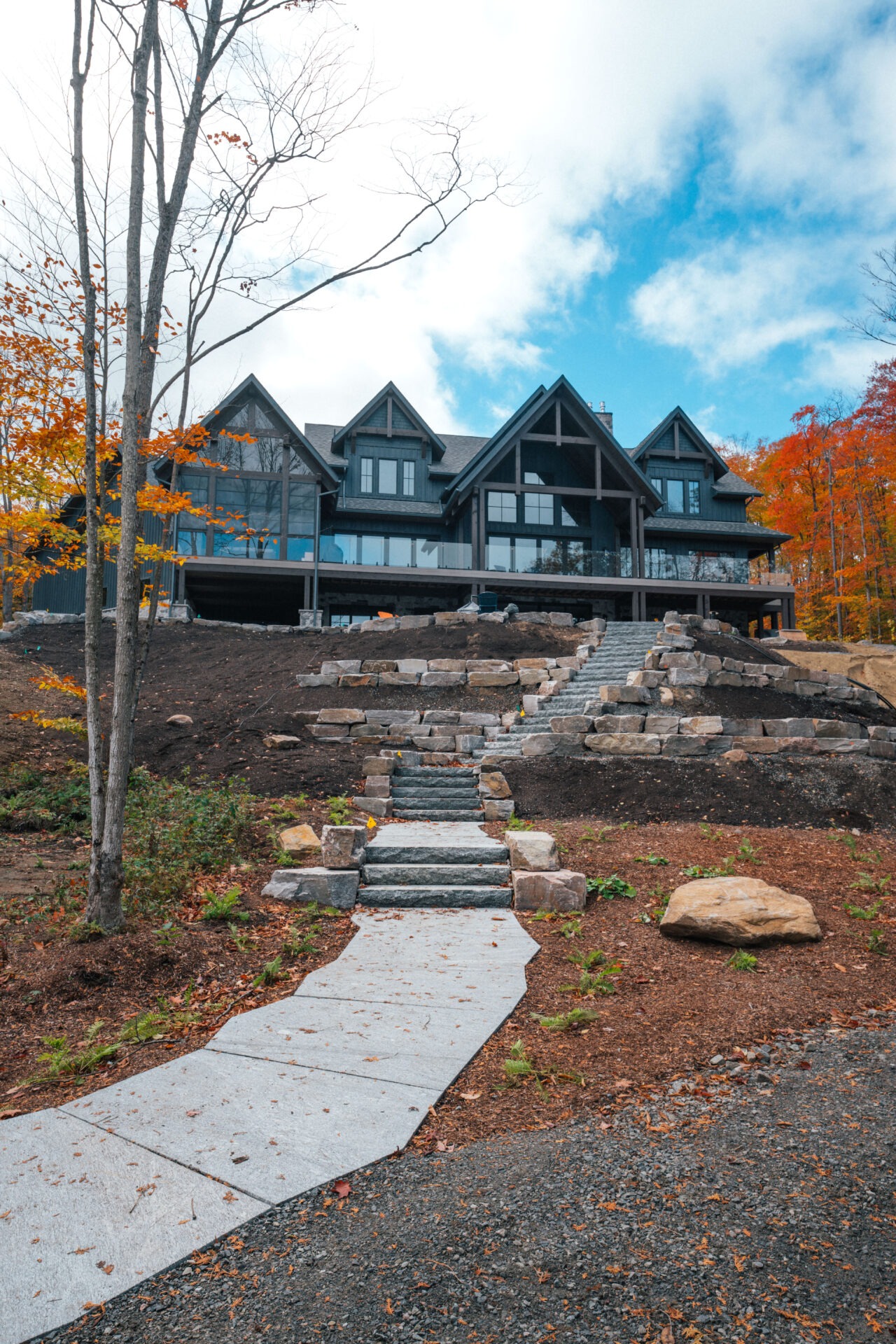 A large contemporary house with multiple gables, stone pathway, autumnal trees, and a bright sky above, surrounded by a natural landscape.