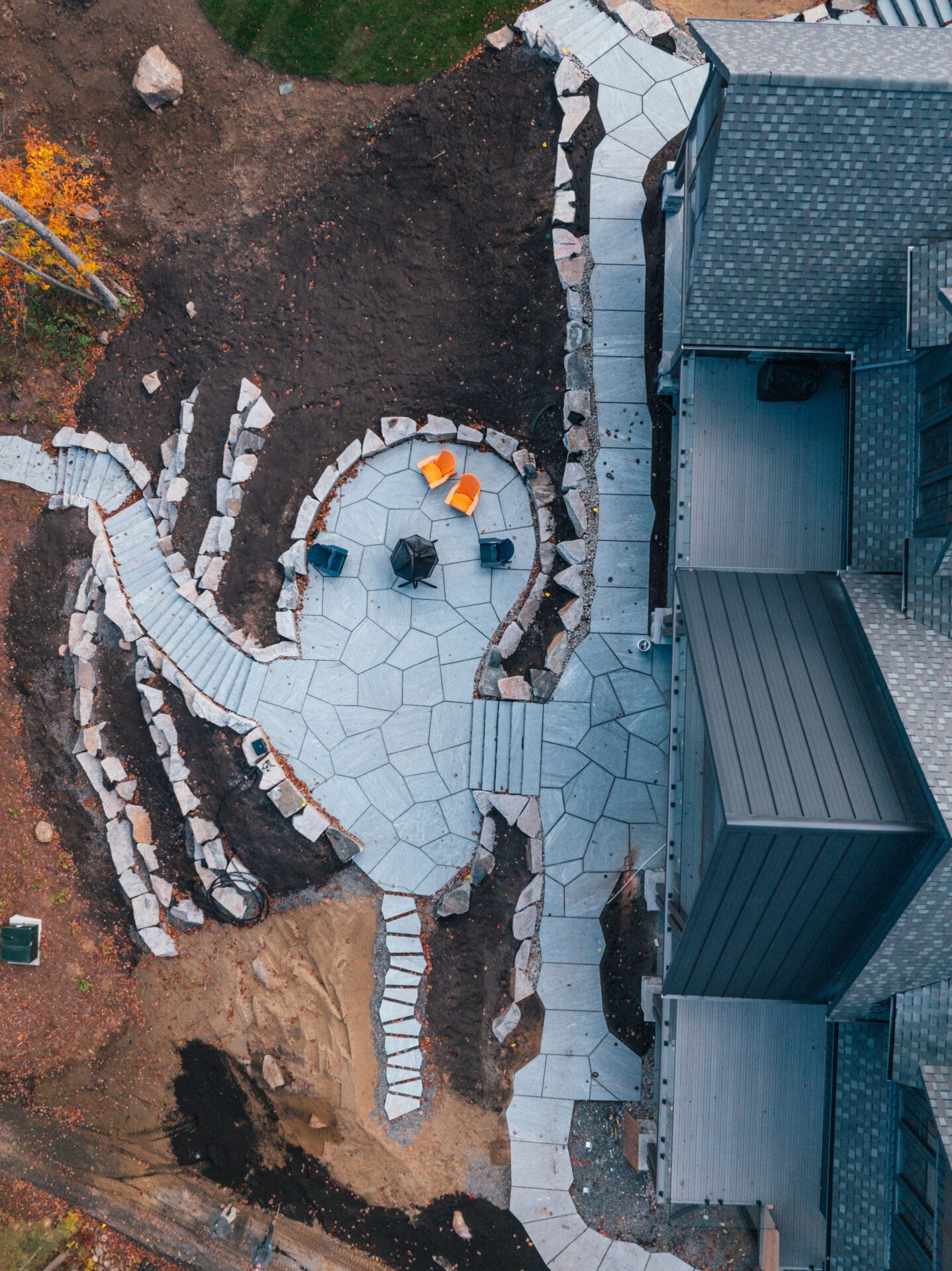 Aerial view of a landscaped backyard featuring stone pathways, surrounded by soil and grass, with colorful chairs arranged on a circular patio.