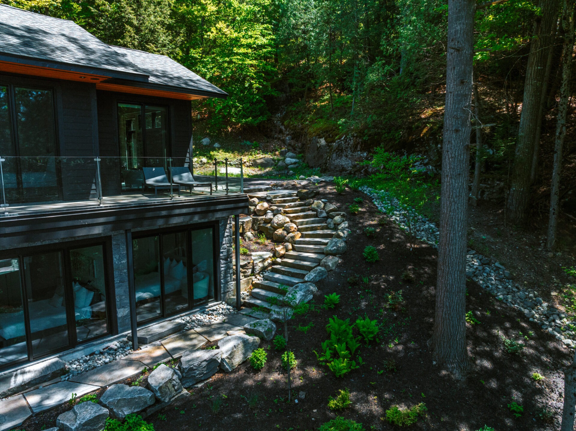 Modern house with large windows next to landscaped stone steps, surrounded by dense forest, under clear blue sky. Peaceful and secluded setting.