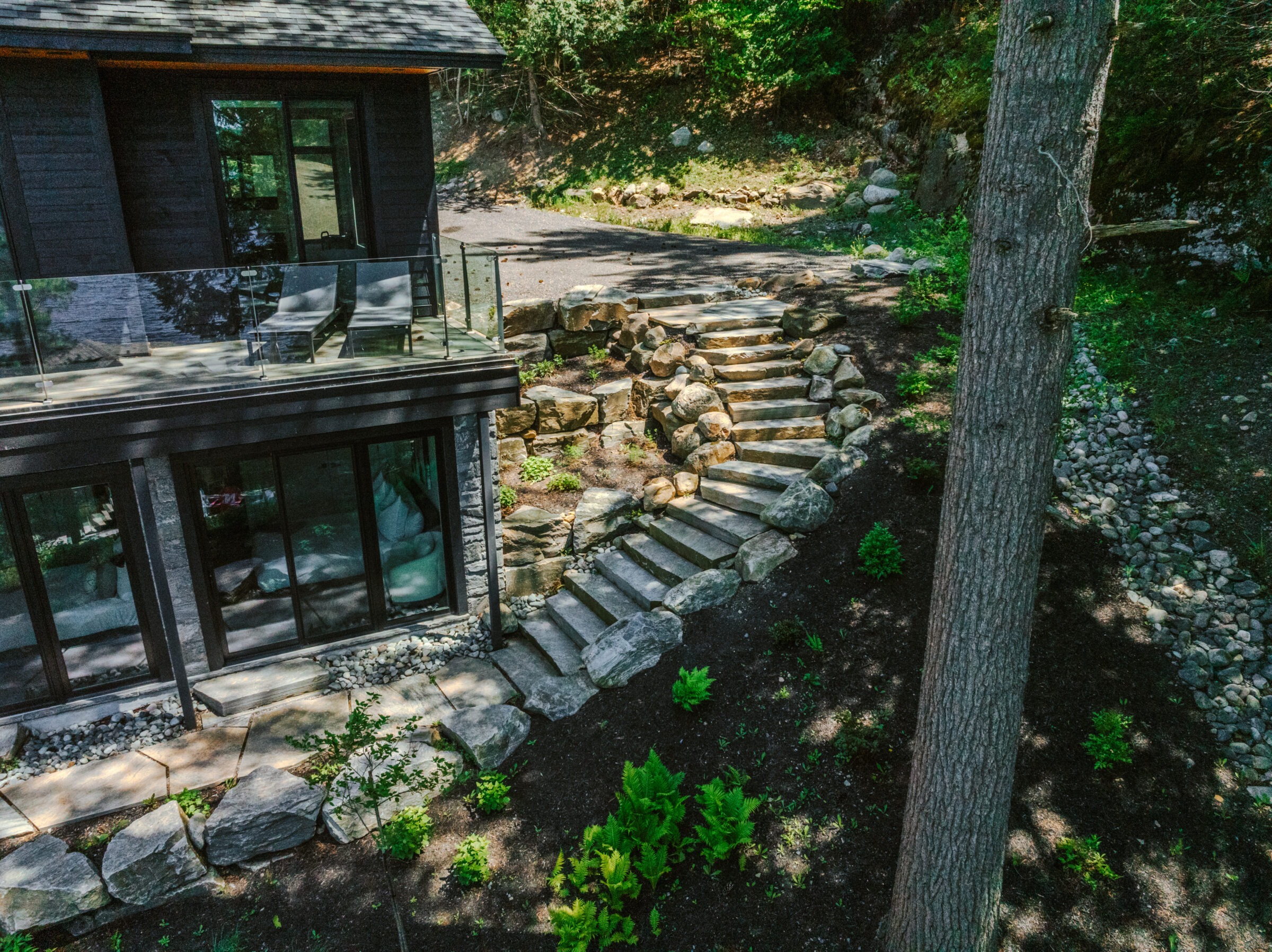 Modern house with glass balcony, stone patio, and stairs leading through a landscaped garden. Surrounded by trees and natural terrain.
