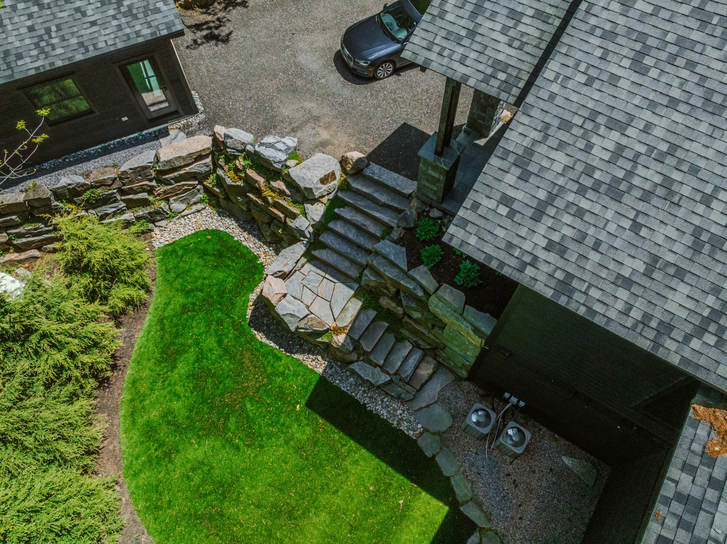 Aerial view of a backyard featuring stone steps, lush green grass, and a parked car beside a house with a shingled roof.