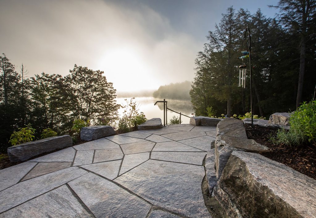 Stone patio with geometric pattern overlooks a serene lake surrounded by trees, illuminated by the rising sun, creating a peaceful atmosphere.
