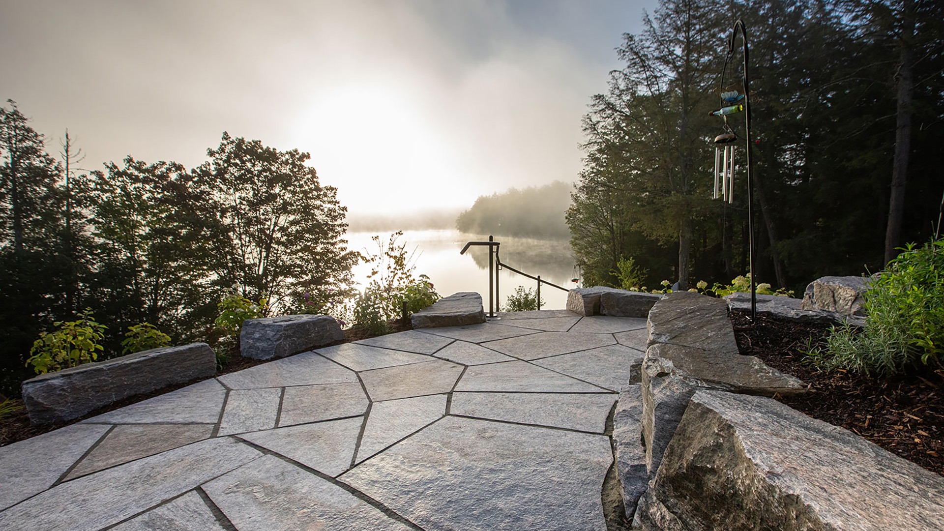 Sunrise over a misty lake, viewed from a stone patio surrounded by trees and plants, with a wind chime hanging nearby.