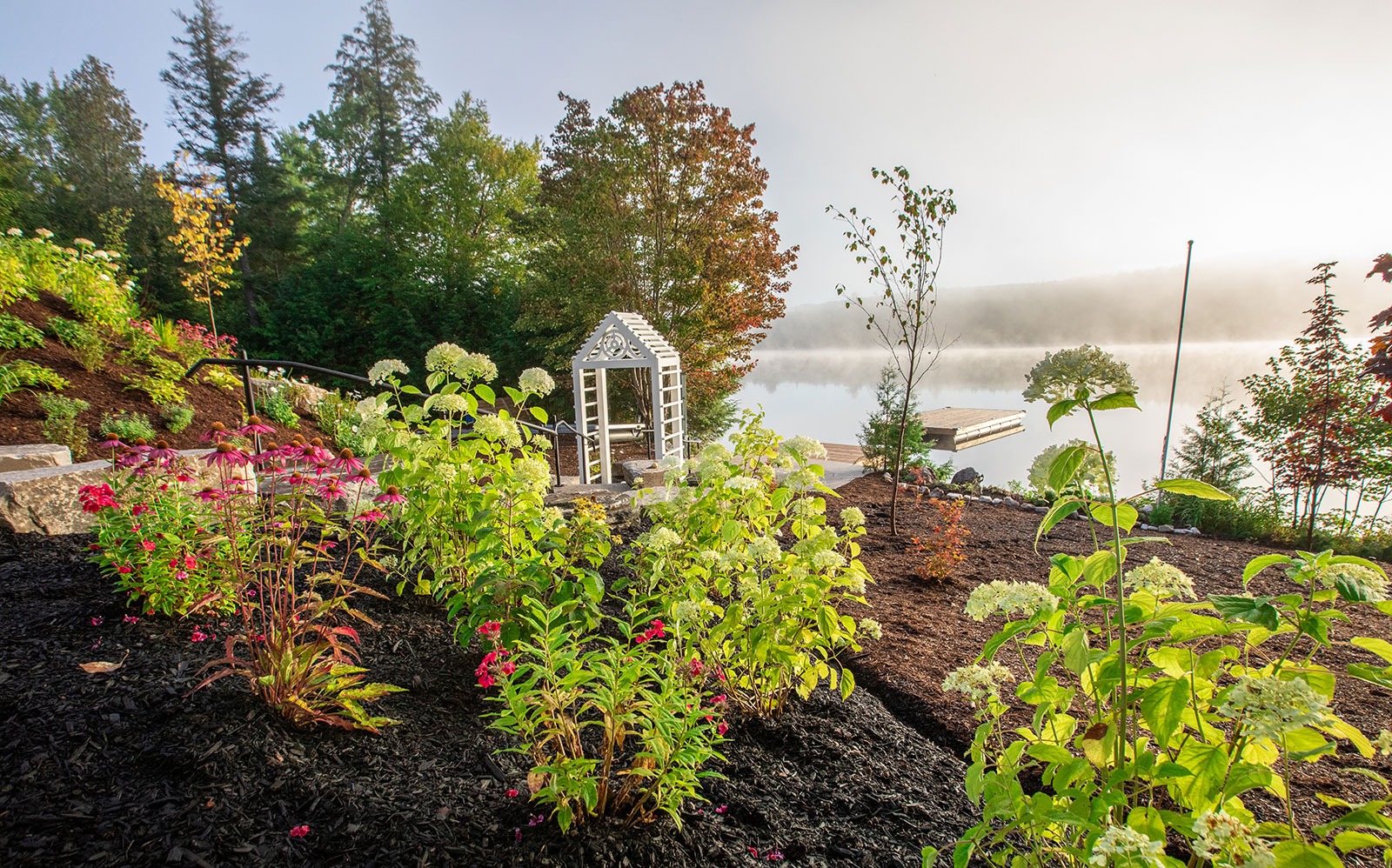 A garden with vibrant flowers and a white trellis overlooks a serene lake surrounded by trees under a misty sky.
