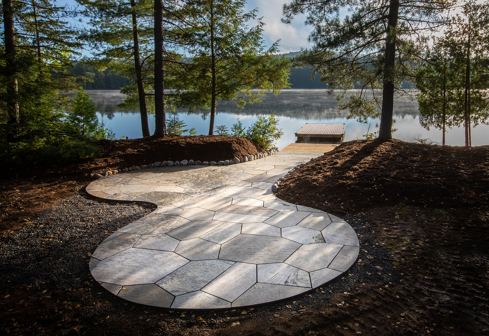 A peaceful lakeside scene with a stone pathway leading to a wooden dock, surrounded by tall trees and serene, reflective water.