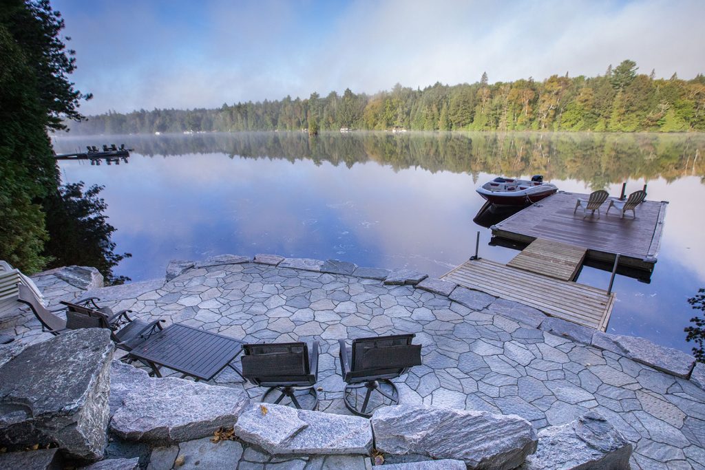 A tranquil lake scene with chairs, a stone patio, dock, and boat. Evergreen trees reflect on calm water under a clear, blue sky.