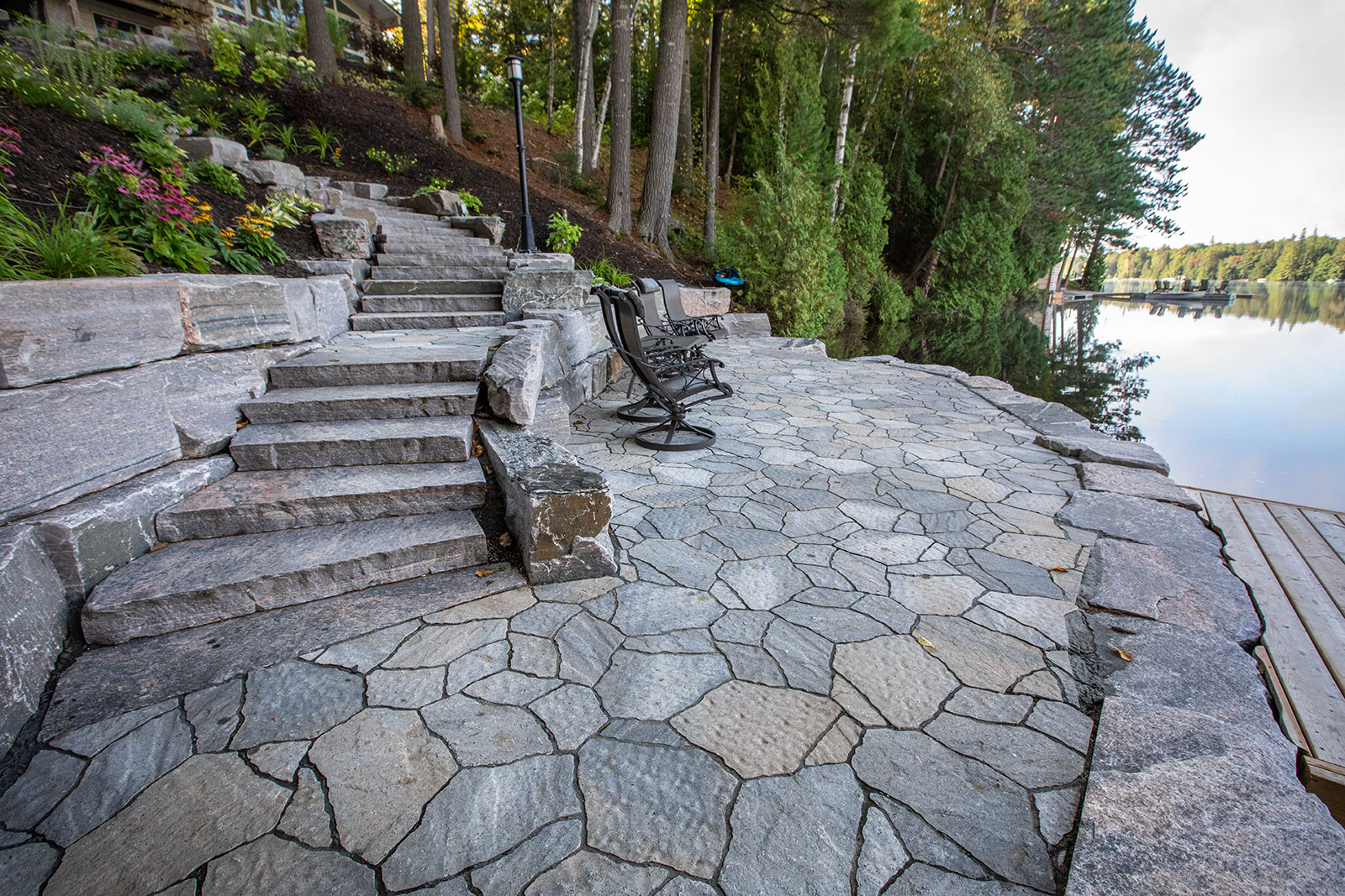 Stone patio with steps, overlooking a calm lake surrounded by trees. Metal chairs are arranged on the patio near the water.
