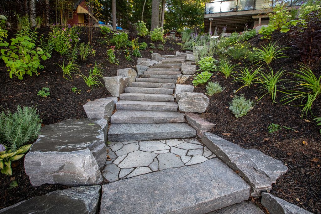 Stone steps ascend through a landscaped garden with lush greenery, surrounded by mulch and rocks, leading toward a house partially visible above.