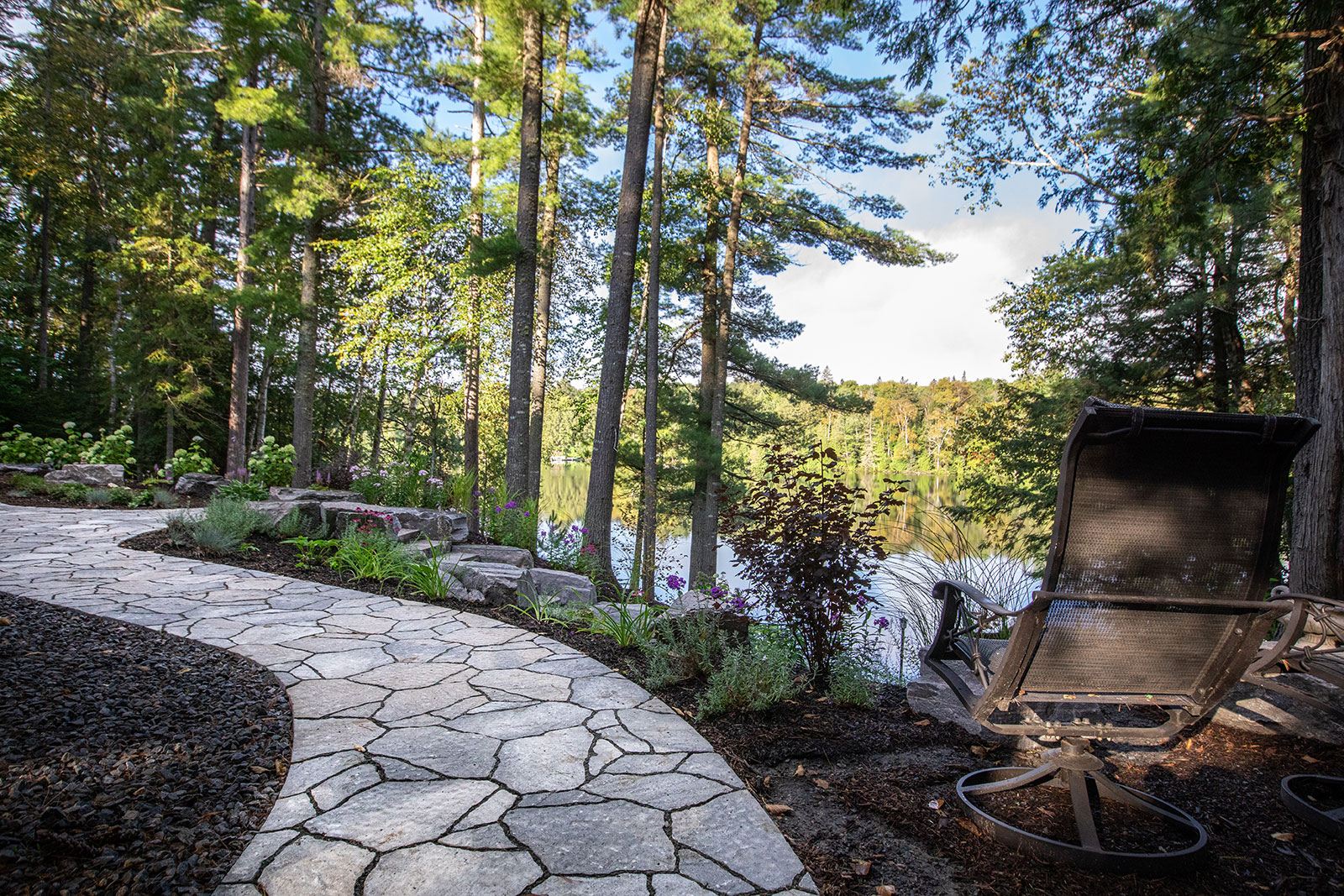 A serene lakeside scene features a stone path, lush trees, and an empty lounge chair, set against a peaceful forest backdrop.