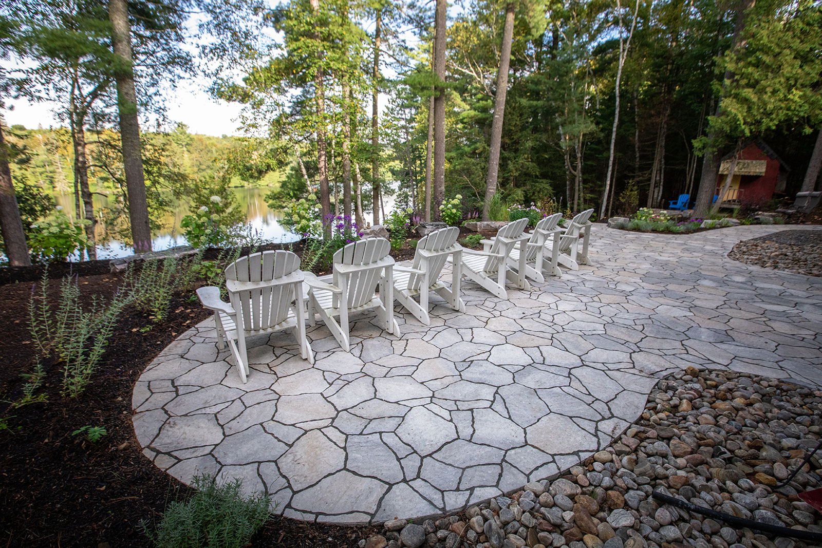 Stone patio with white chairs faces a tranquil lake, surrounded by lush trees and landscaped garden, creating a peaceful outdoor setting.