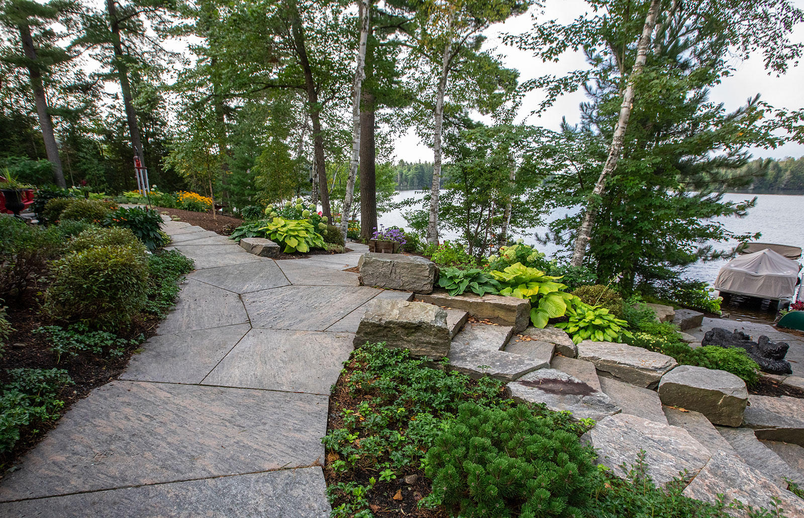 A stone pathway winds through lush, landscaped gardens by a lake, surrounded by trees and a docked boat under a cover.