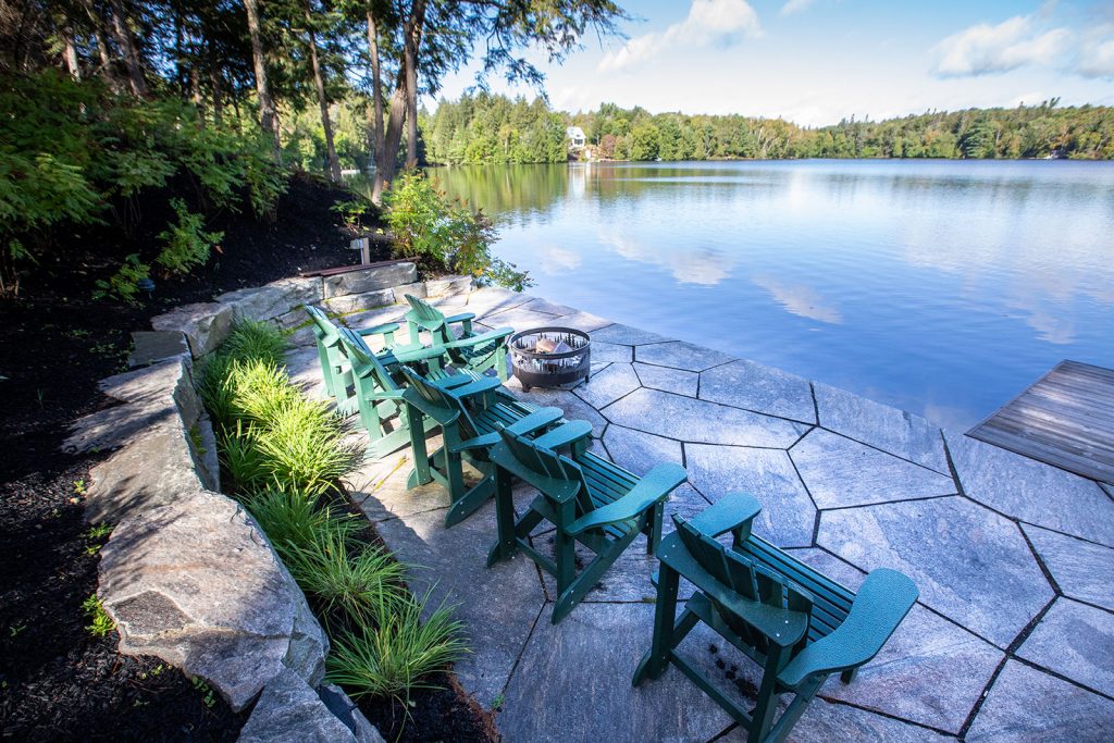 Lakeside patio with green Muskoka chairs, fire pit, and lush trees. Clear sky reflects on the calm water. Peaceful, scenic setting.