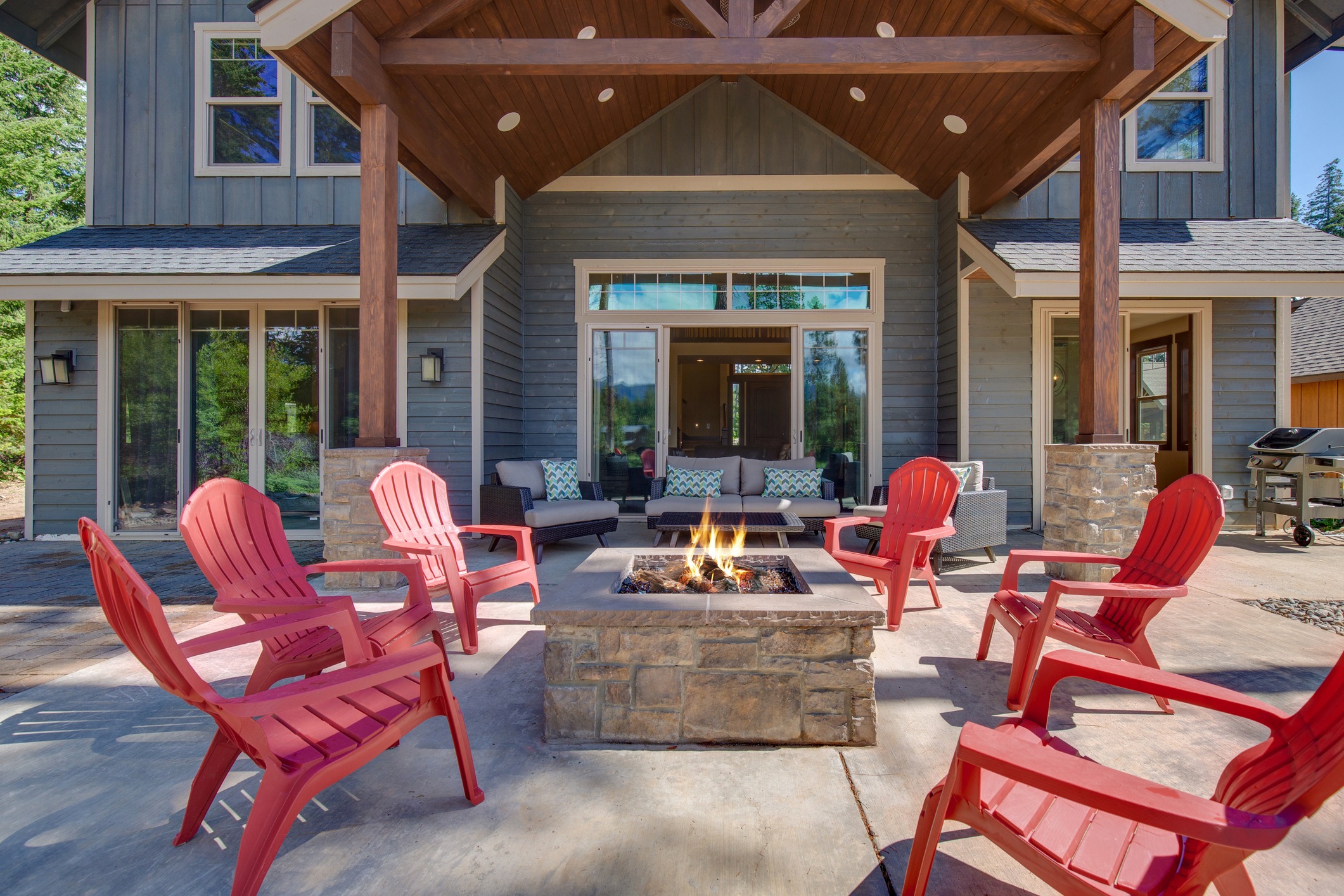 A cozy patio features red chairs surrounding a stone fire pit, set in front of a modern wooden house with large windows.
