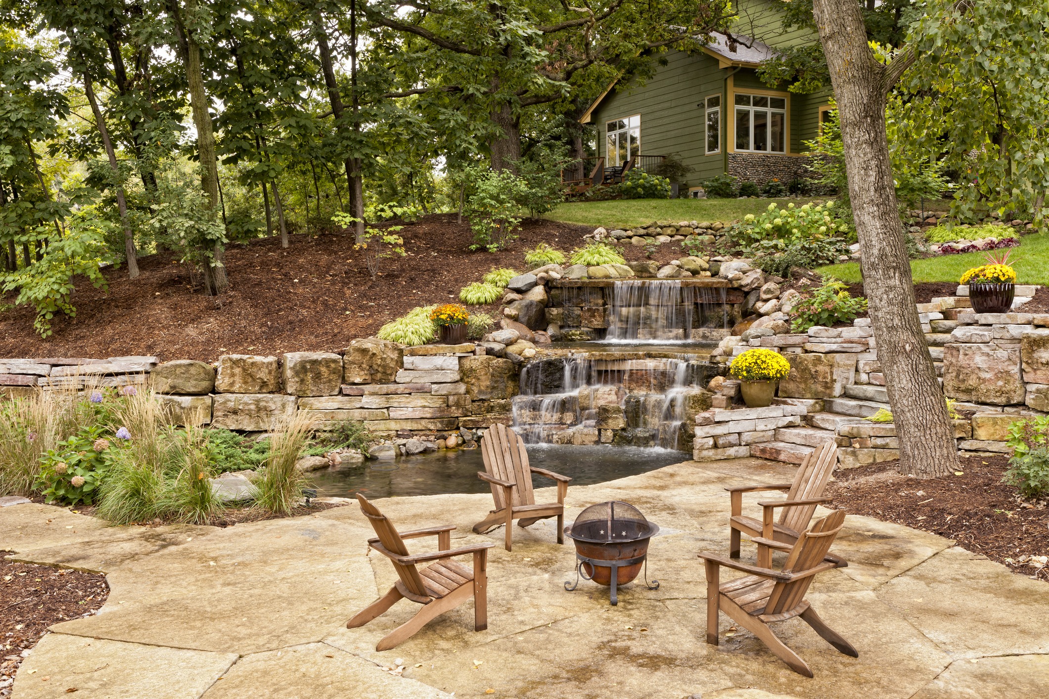 Four wooden chairs surround a fire pit on a stone patio, overlooking a cascading waterfall and a lush garden with a charming house nearby.