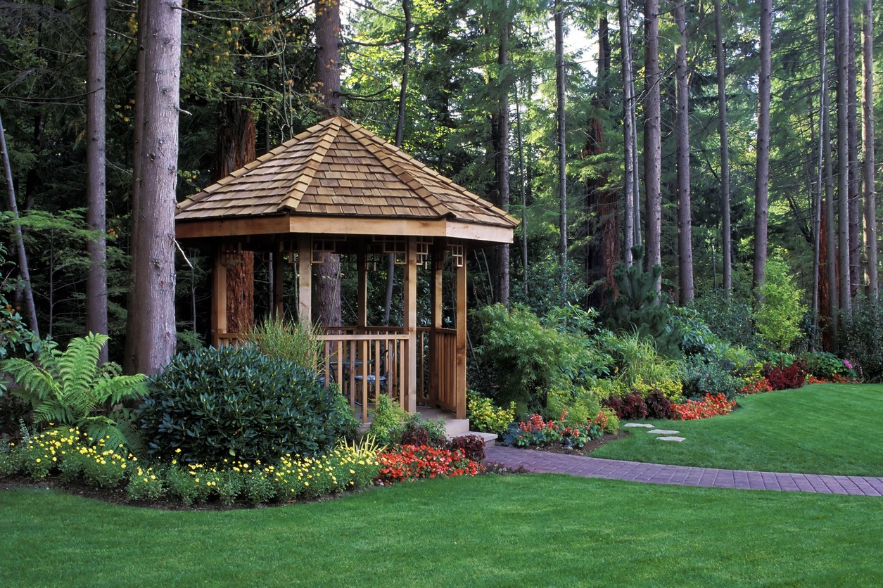 A wooden gazebo stands among tall trees and colorful flower beds, surrounded by lush green grass and a brick pathway in a forest setting.
