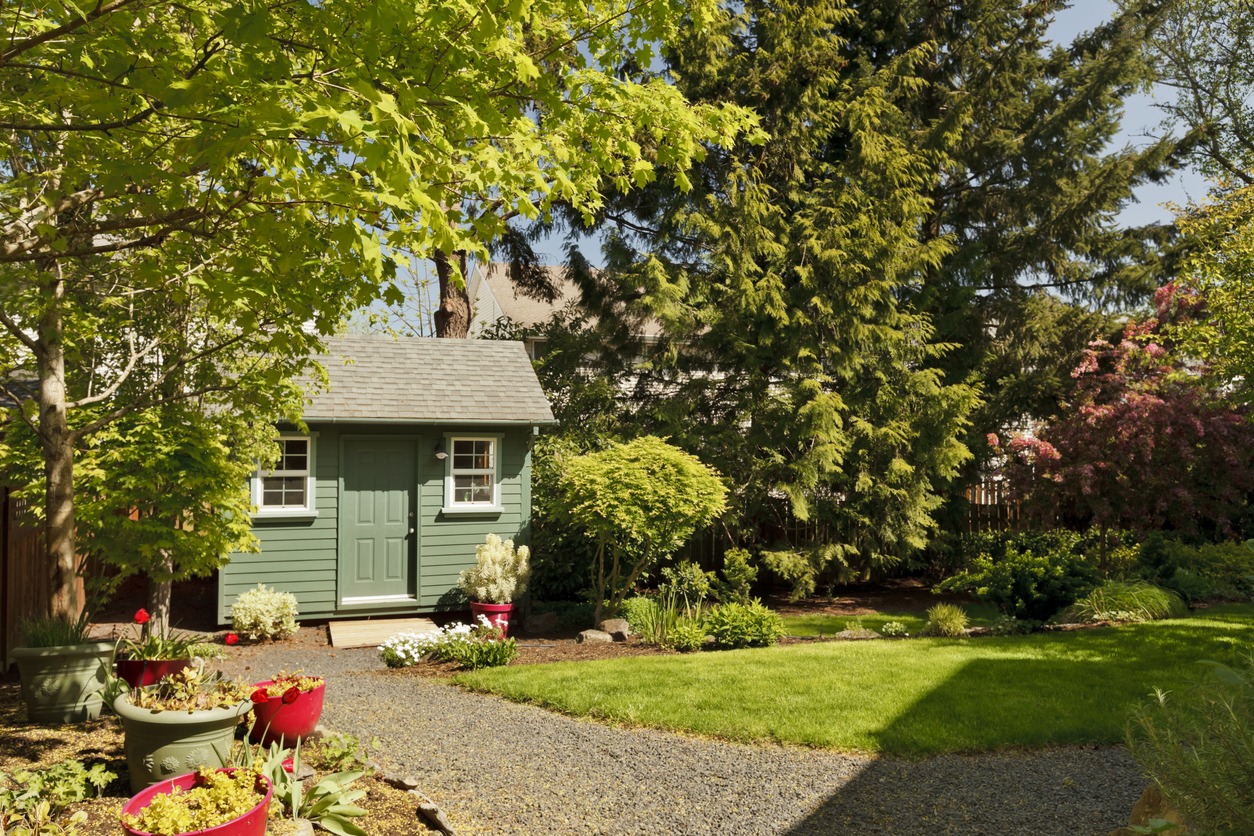 A small green garden shed surrounded by lush trees, potted plants, and a stone path in a well-maintained backyard garden.
