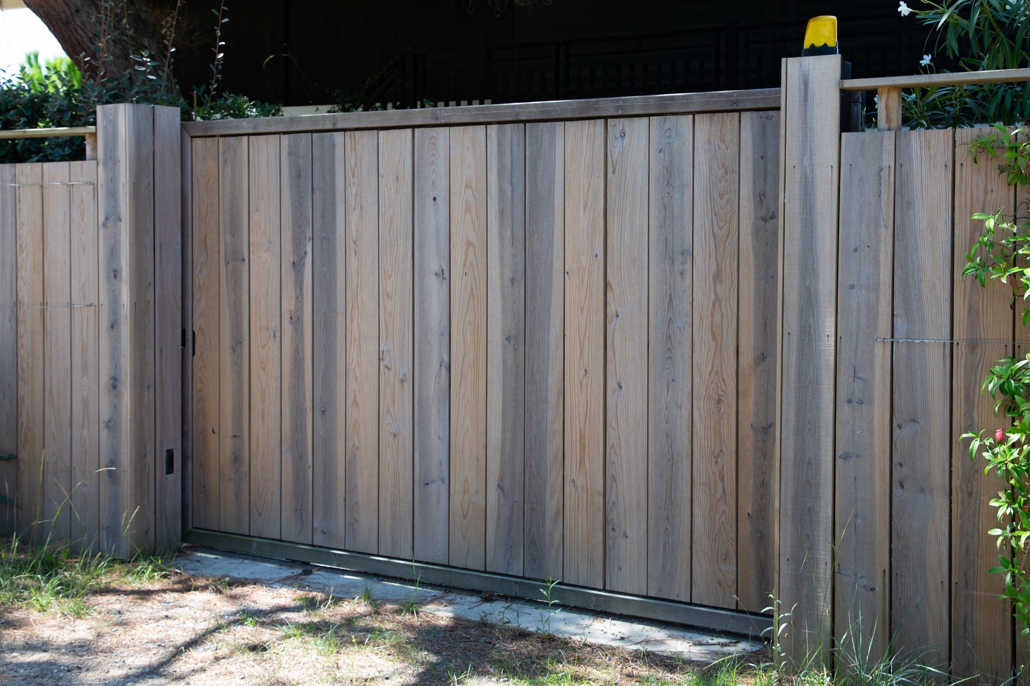 Wooden gate in outdoor setting, surrounded by greenery. The gate has vertical slats and a yellow light fixture on top, casting shadows nearby.