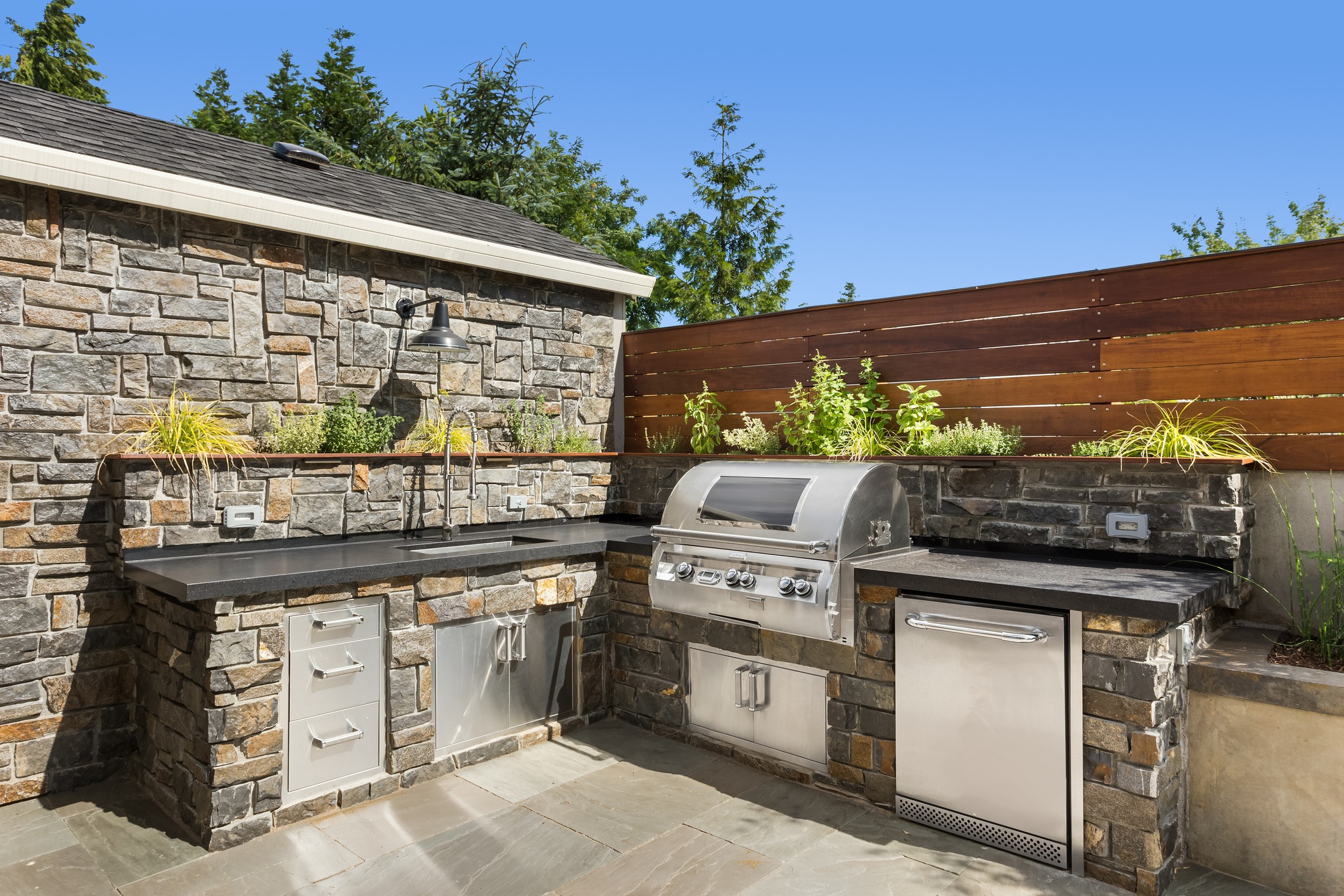 Outdoor kitchen with stone walls, stainless steel grill, modern appliances, sink, and planters under clear blue sky; wooden fence and greenery surrounding.